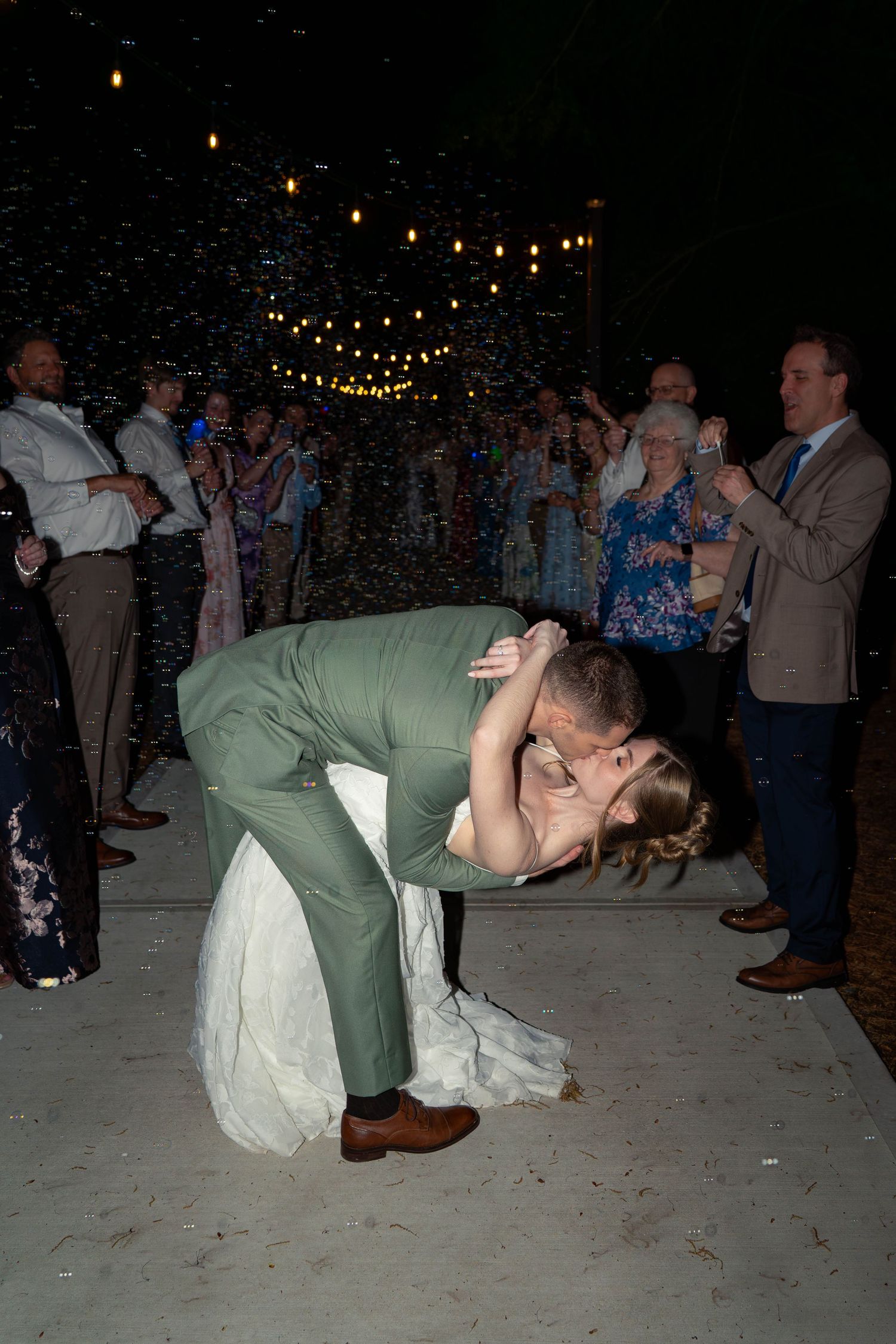 Groom in green suit dips bride in white gown during first dance with confetti falling.