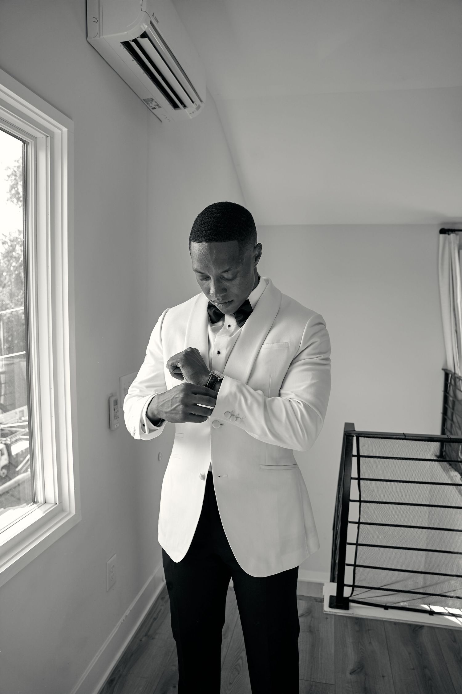 Groom in white tuxedo jacket fastening cufflinks while getting ready in black and white photo.