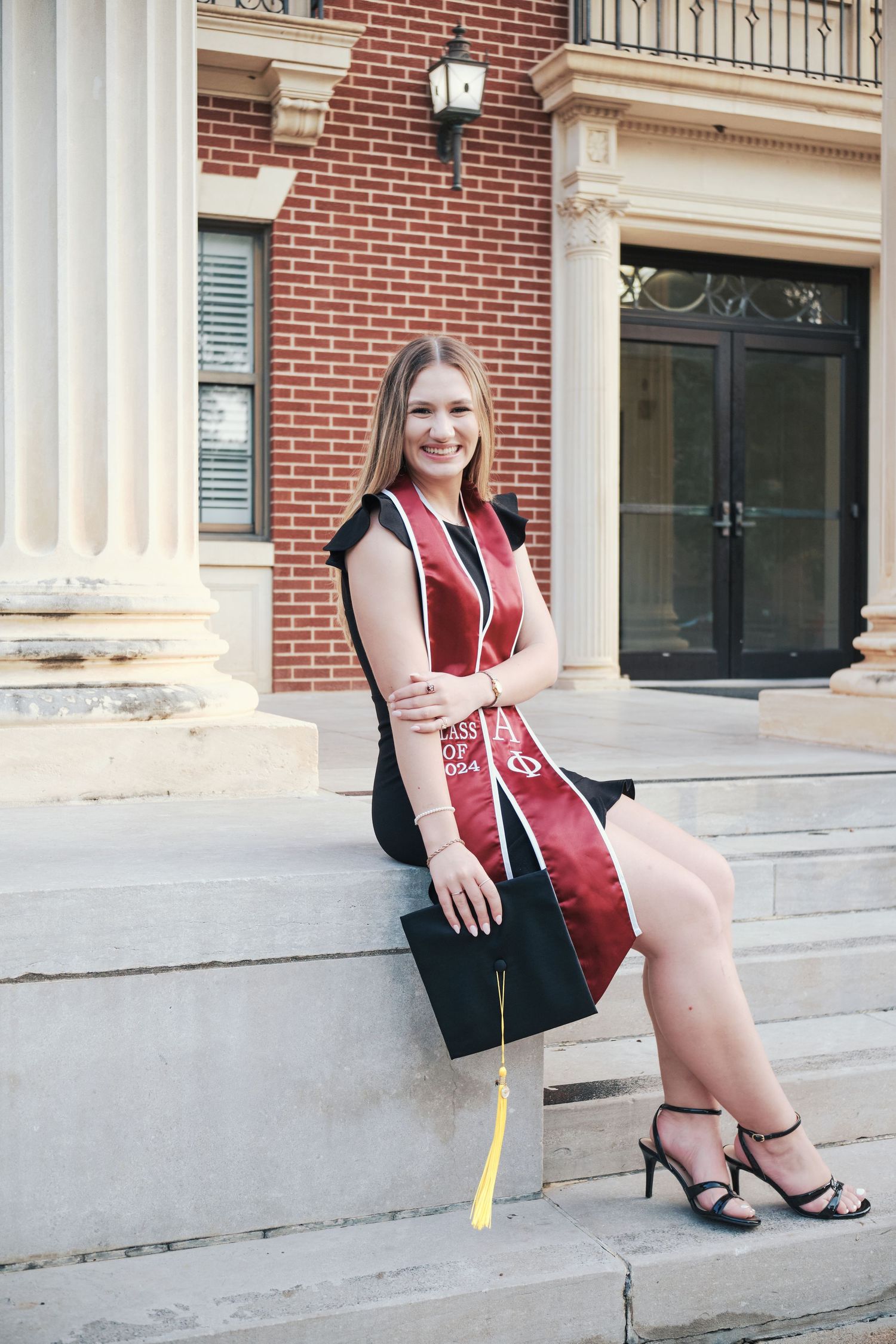 Graduate in black dress and maroon stole sits on steps holding cap at brick university building. North Carolina Photographer.
