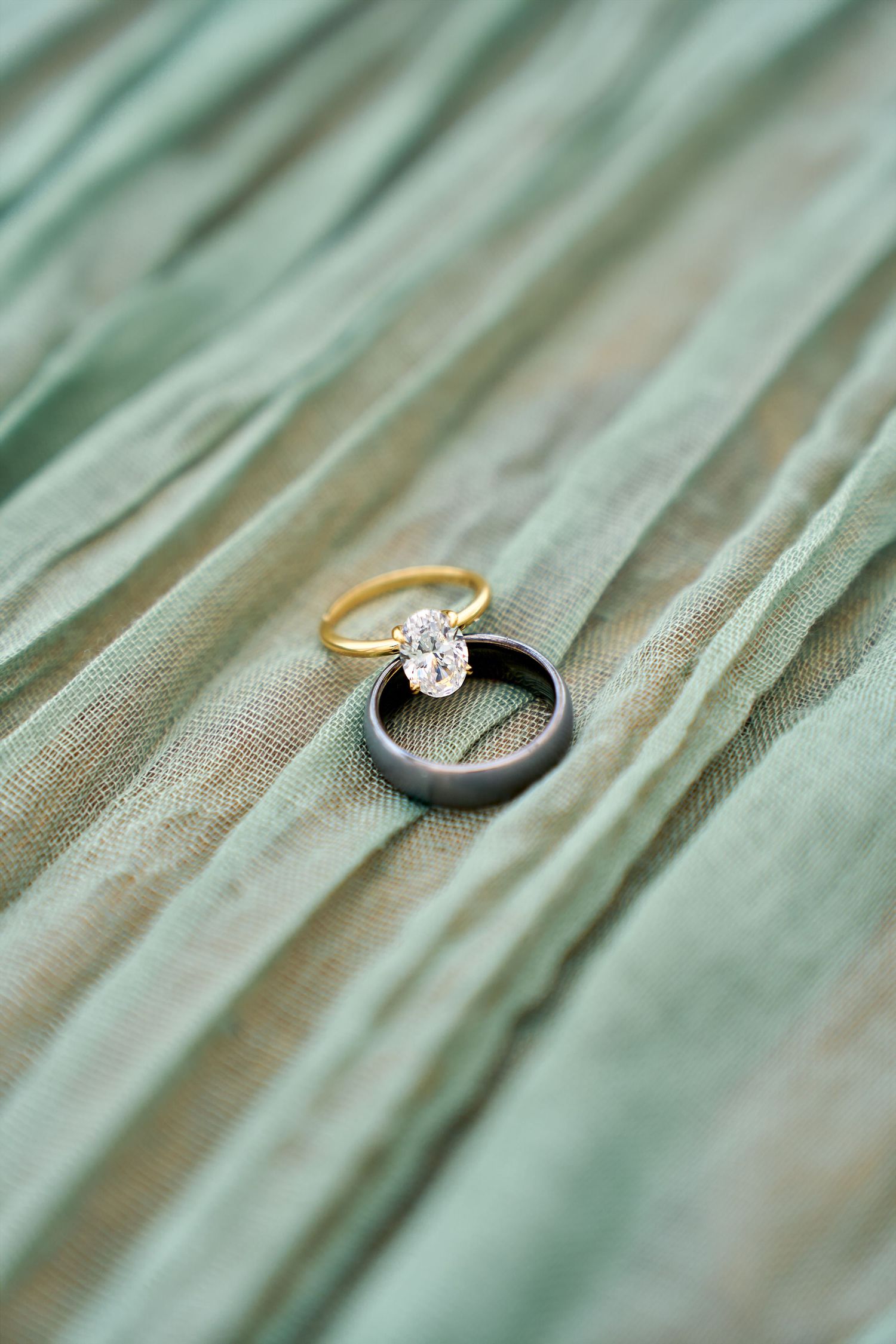 Wedding rings with gold engagement ring and black band on soft sage green fabric with shallow depth of field.