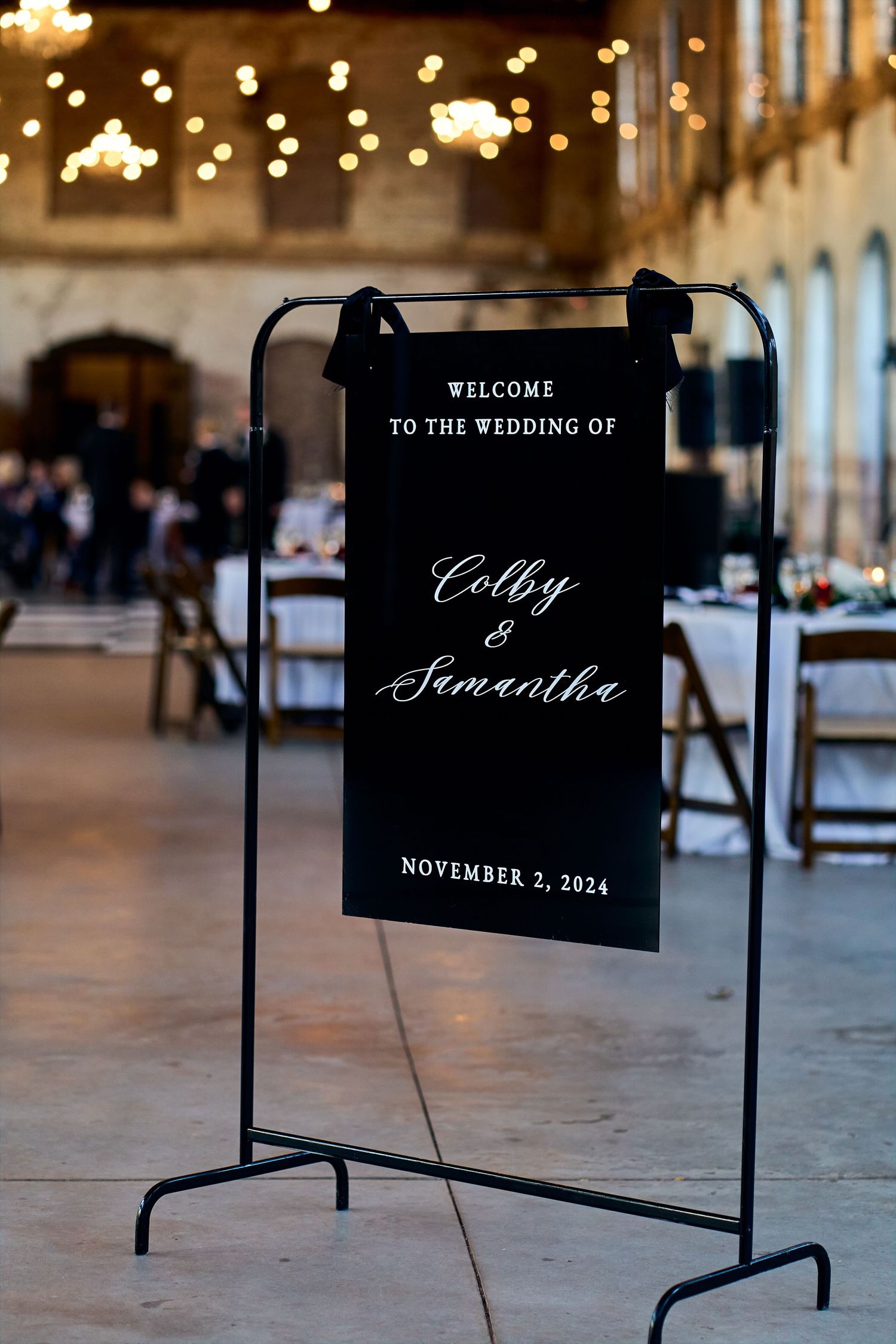 Black welcome sign on metal stand displays wedding details with string lights bokeh in rustic indoor venue background.