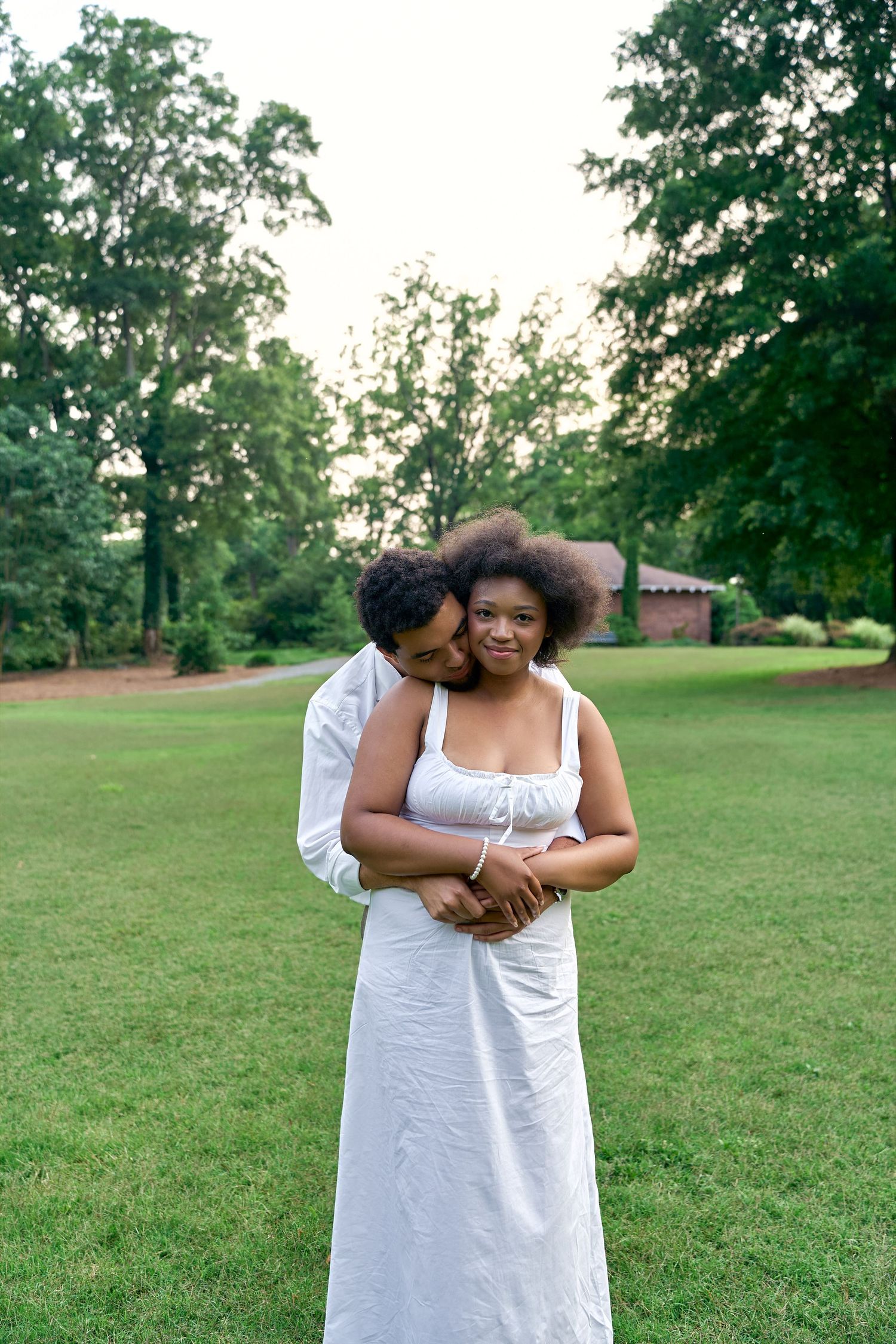 Two people in white dresses embrace while standing together on a grassy lawn in a park setting.