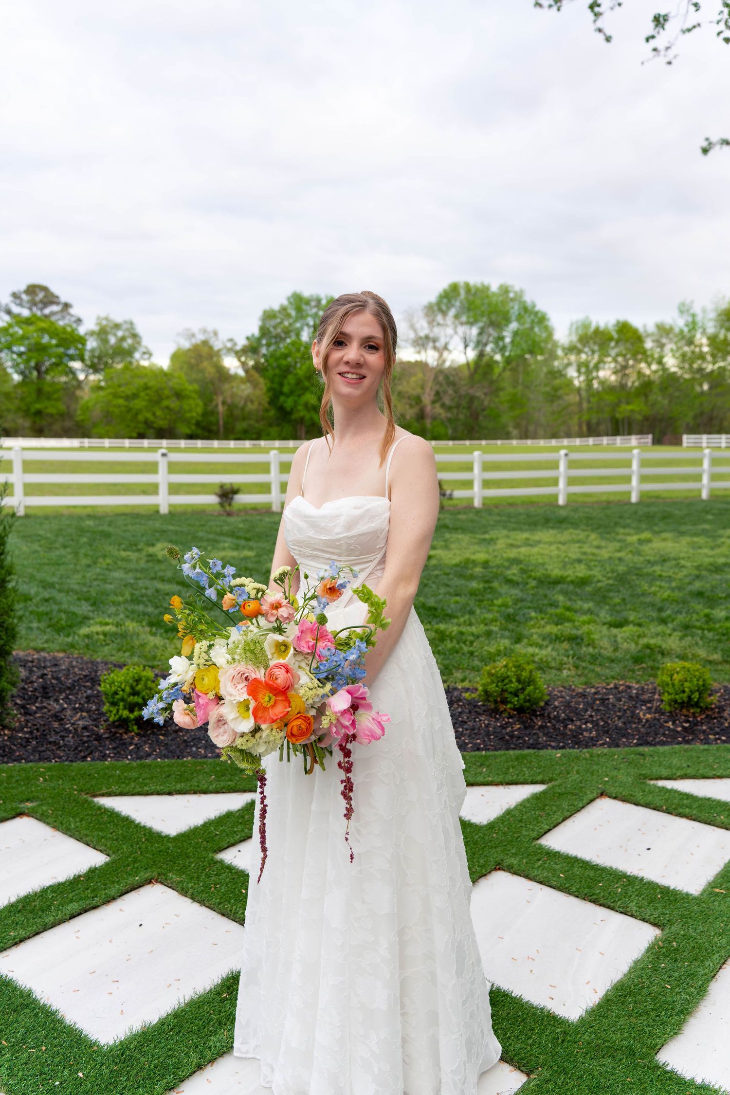 Smiling bride in strapless white lace gown holding vibrant colorful bouquet at outdoor venue.