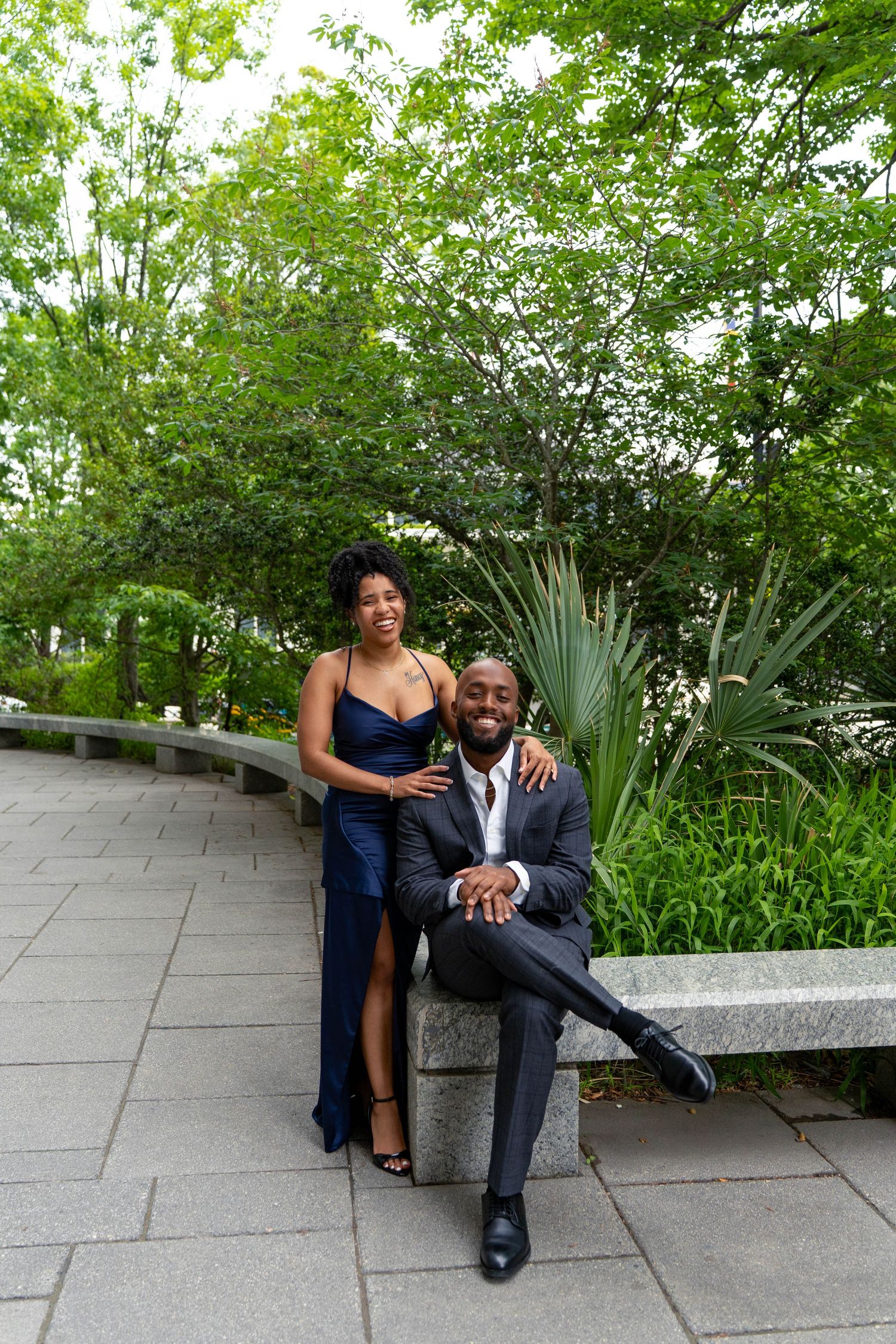 Smiling Black couple posing in a lush garden, woman in navy gown standing beside seated man in suit.