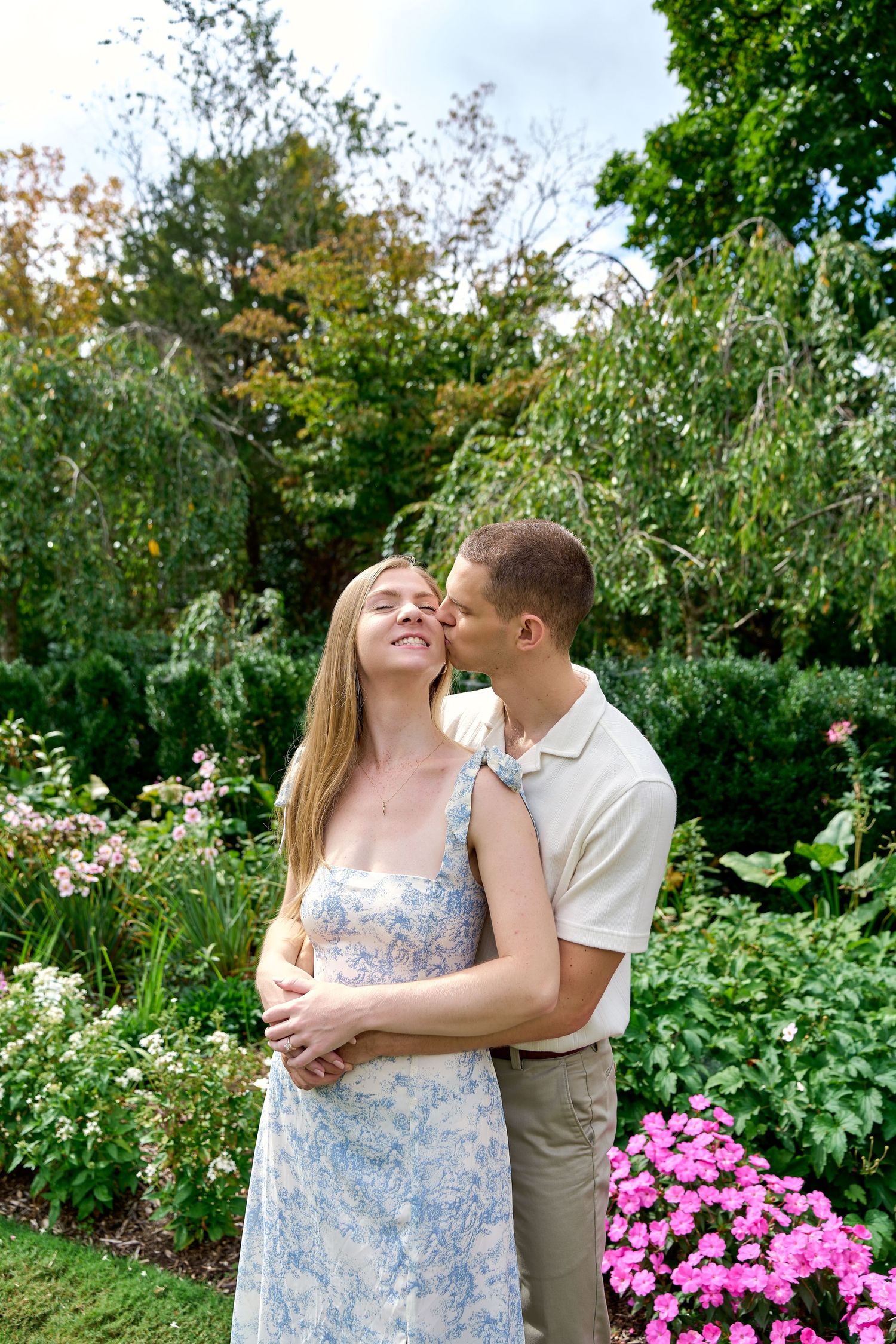 Man tenderly kisses woman on cheek while embracing her in a blooming garden with pink flowers.
