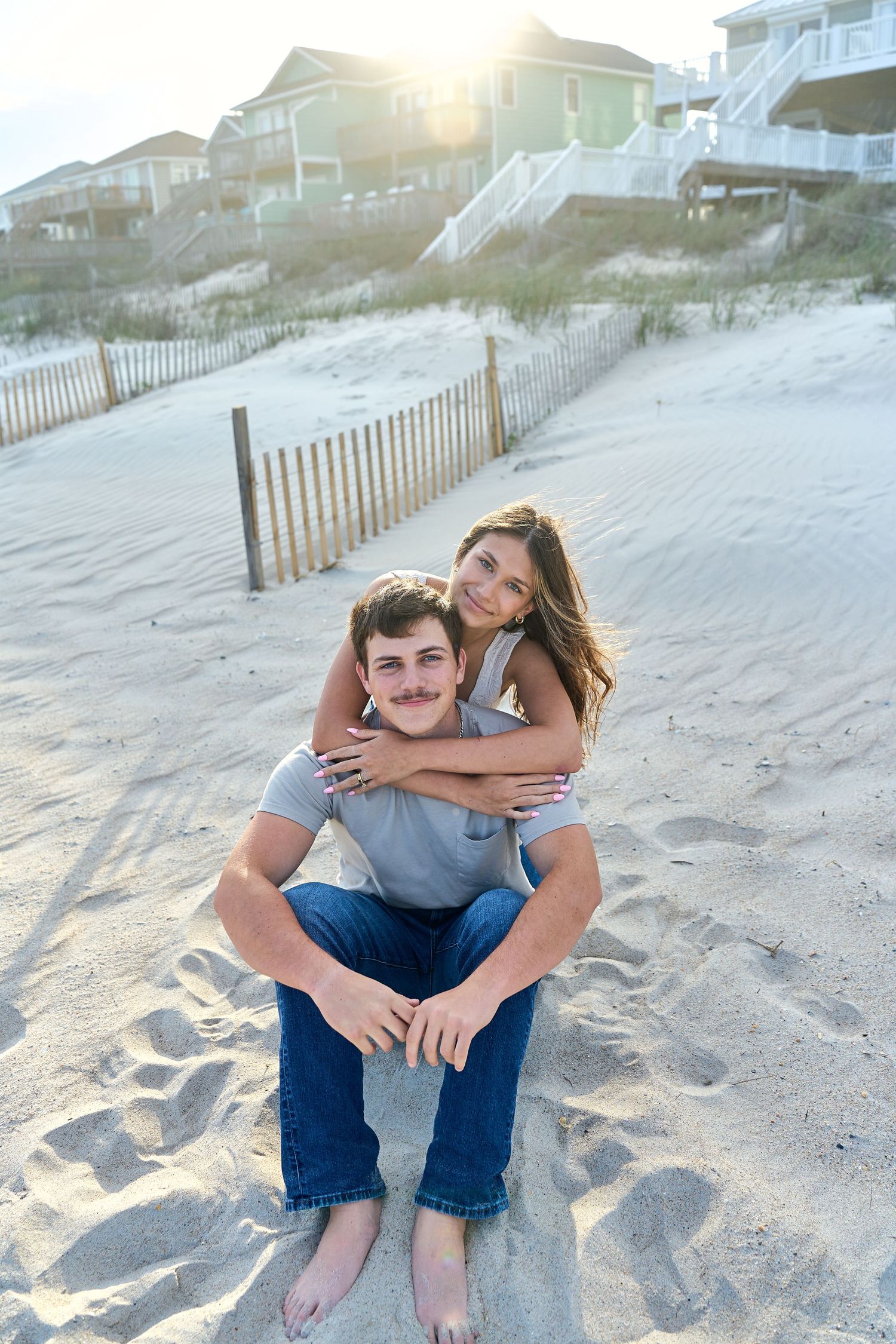 A couple sits together on a sandy beach at sunset with beach houses and dune fences visible in the background.