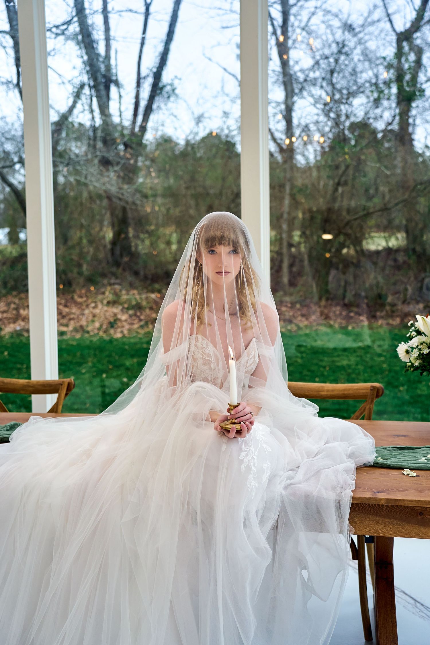 A bride in a flowing white wedding gown and veil sits at a wooden table with a natural outdoor backdrop.