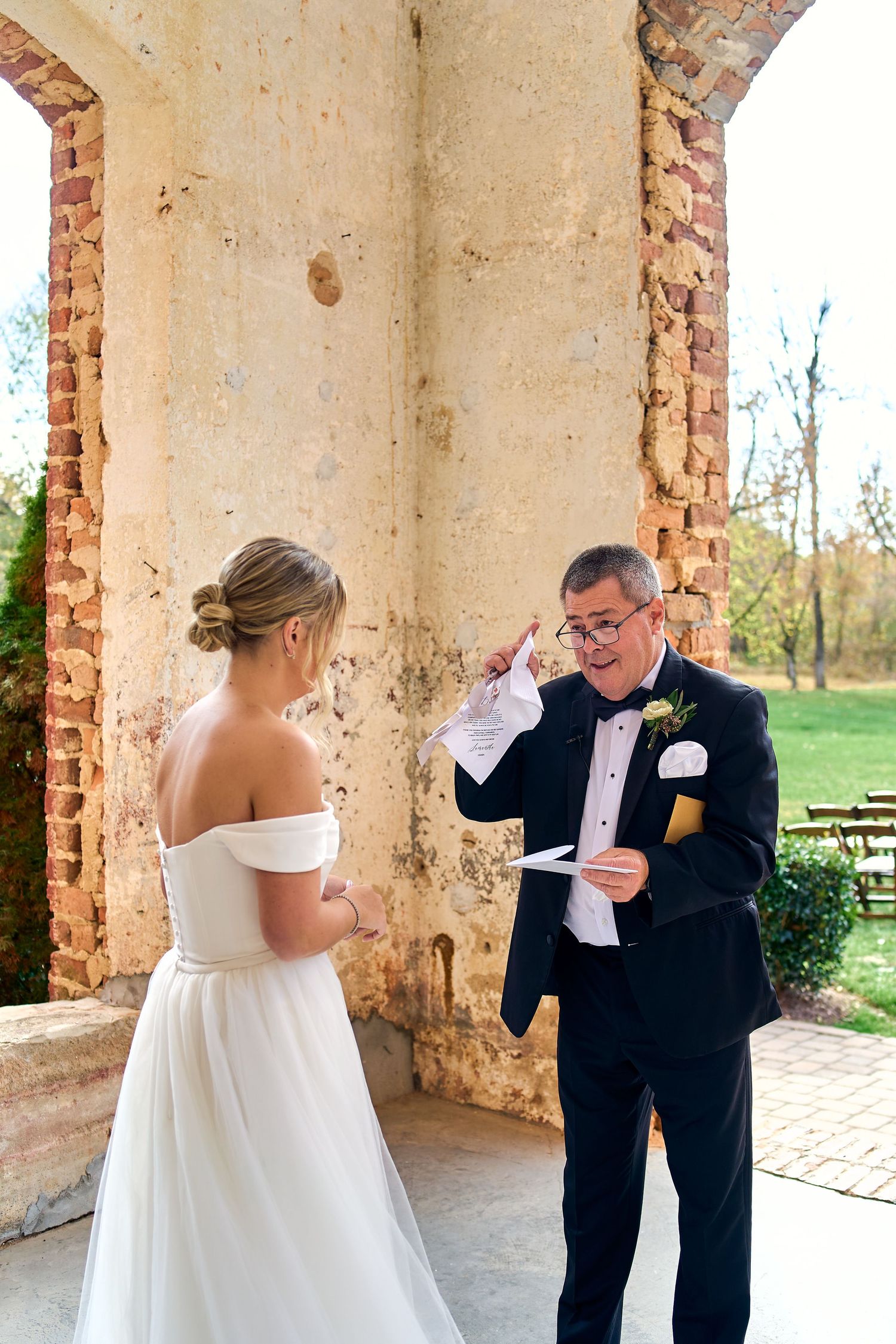 On a sunny day, a person in an off-shoulder white gown exchanges vows with someone in a tuxedo under a stone archway.