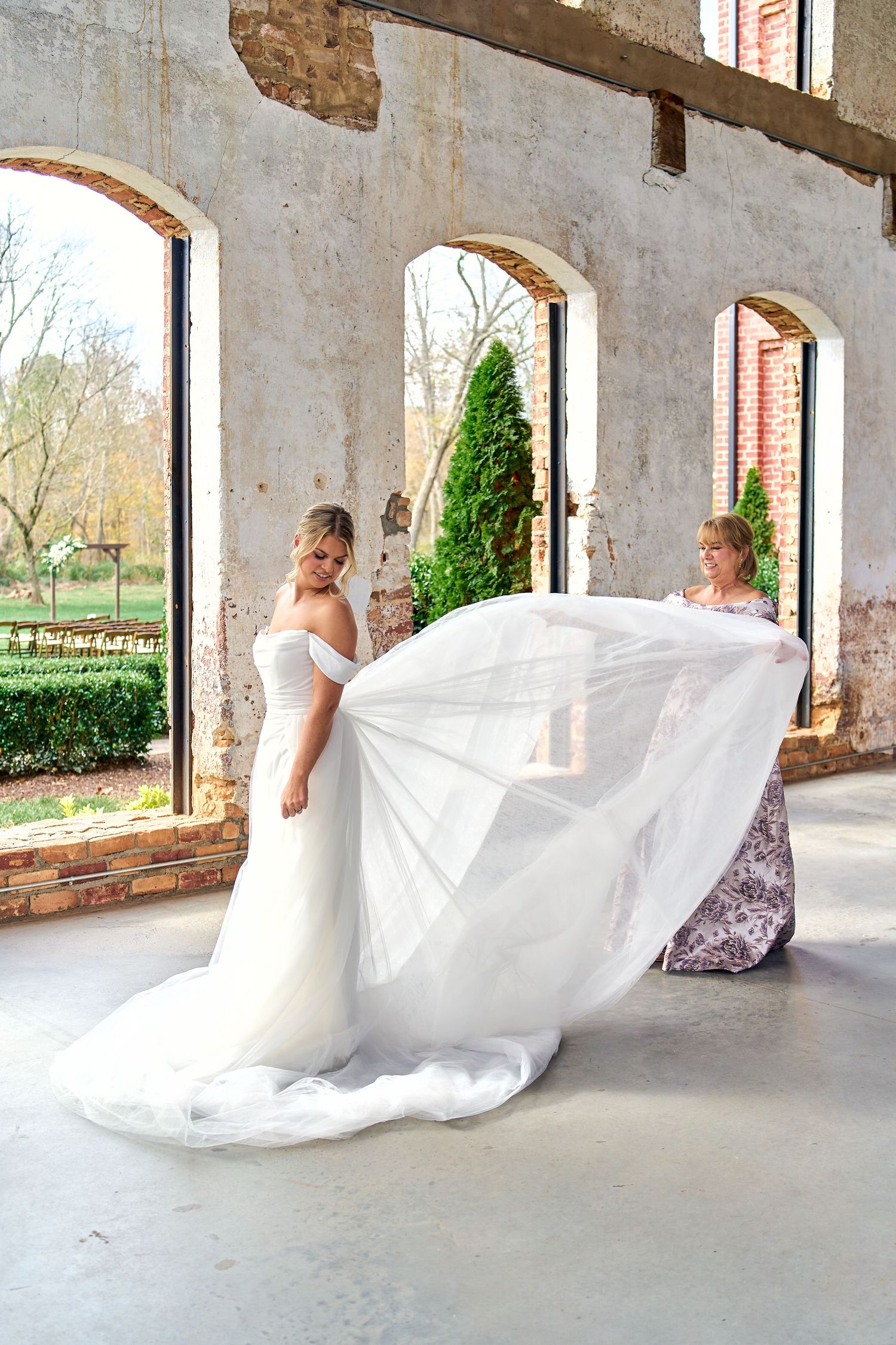 A flowing white wedding gown billows in the breeze within an elegant rustic venue with arched windows and exposed brick walls.