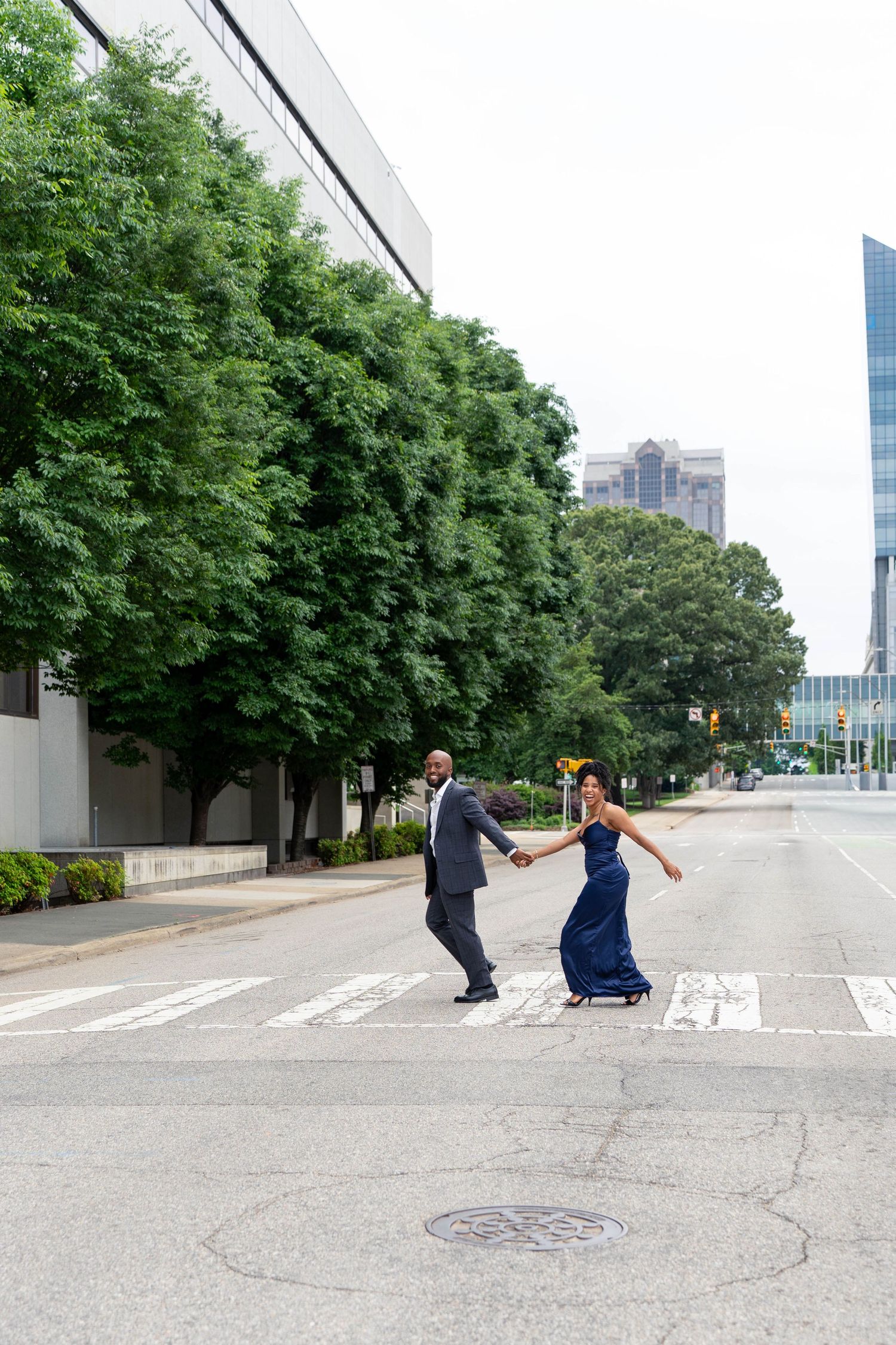 A couple in formal attire holds hands while crossing an empty urban street intersection.