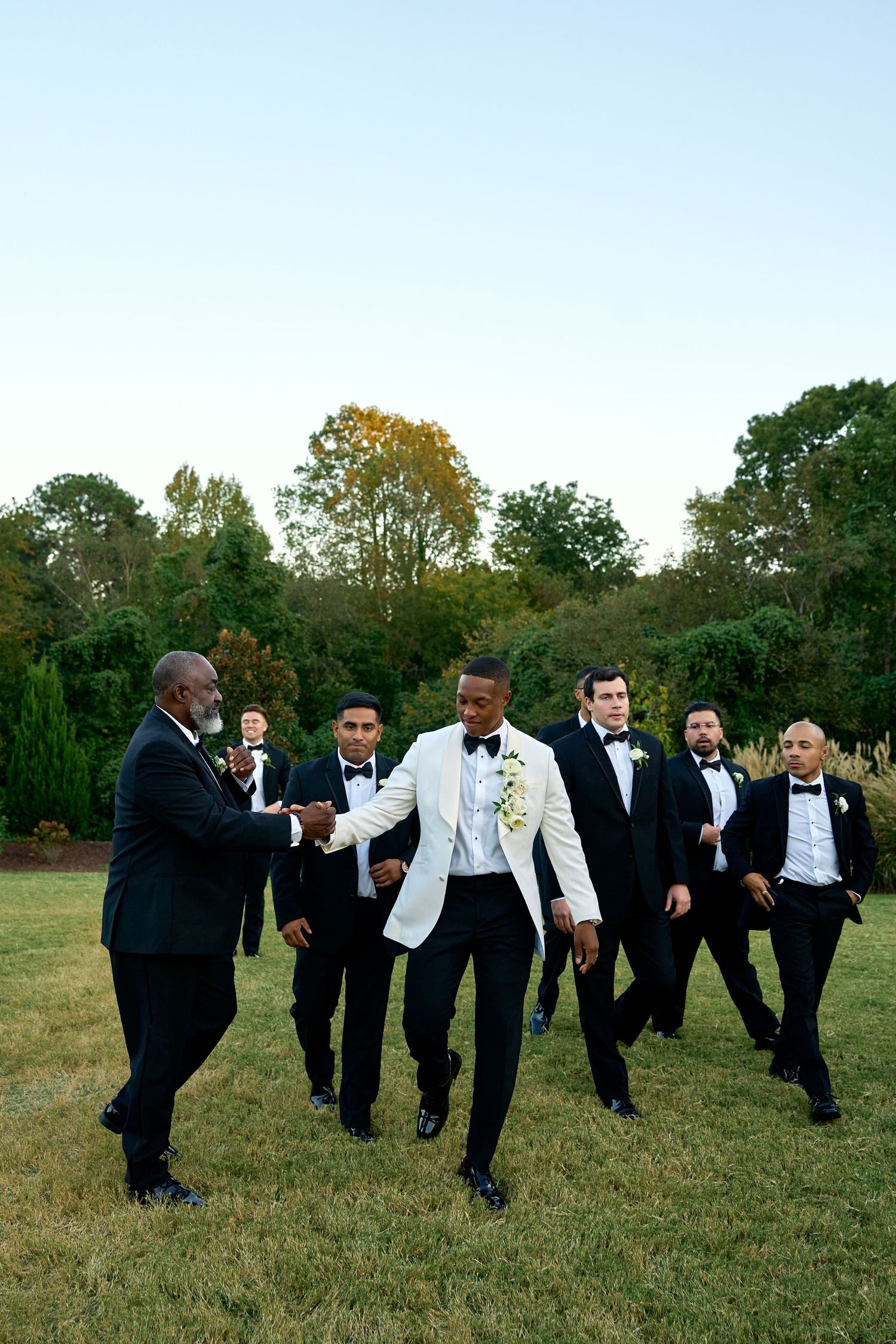 Groom in white tuxedo jacket shakes hands with groomsmen in black tuxedos on green lawn at outdoor wedding.
