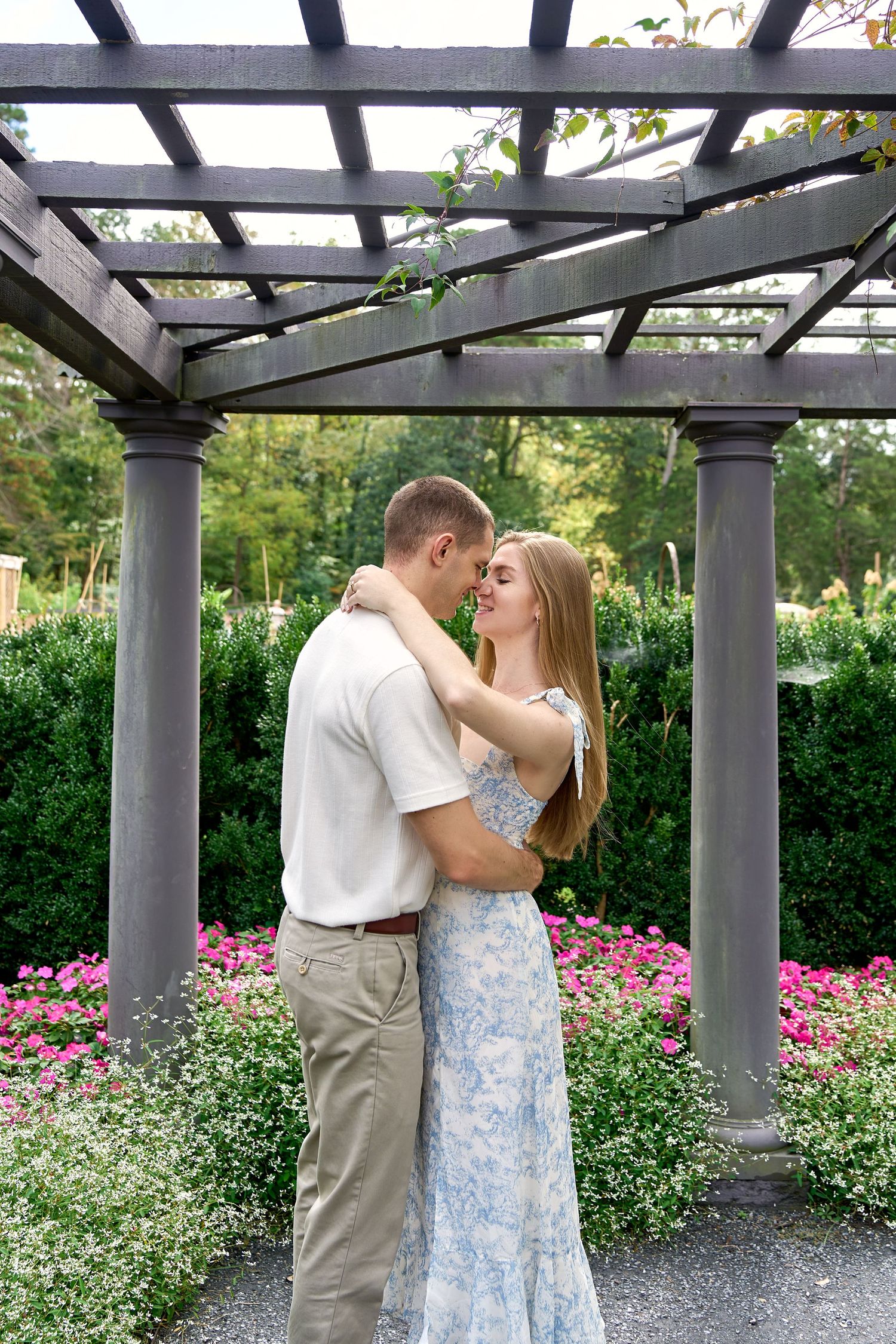 Couple embracing and about to kiss under a rustic wooden pergola with lush green garden backdrop.