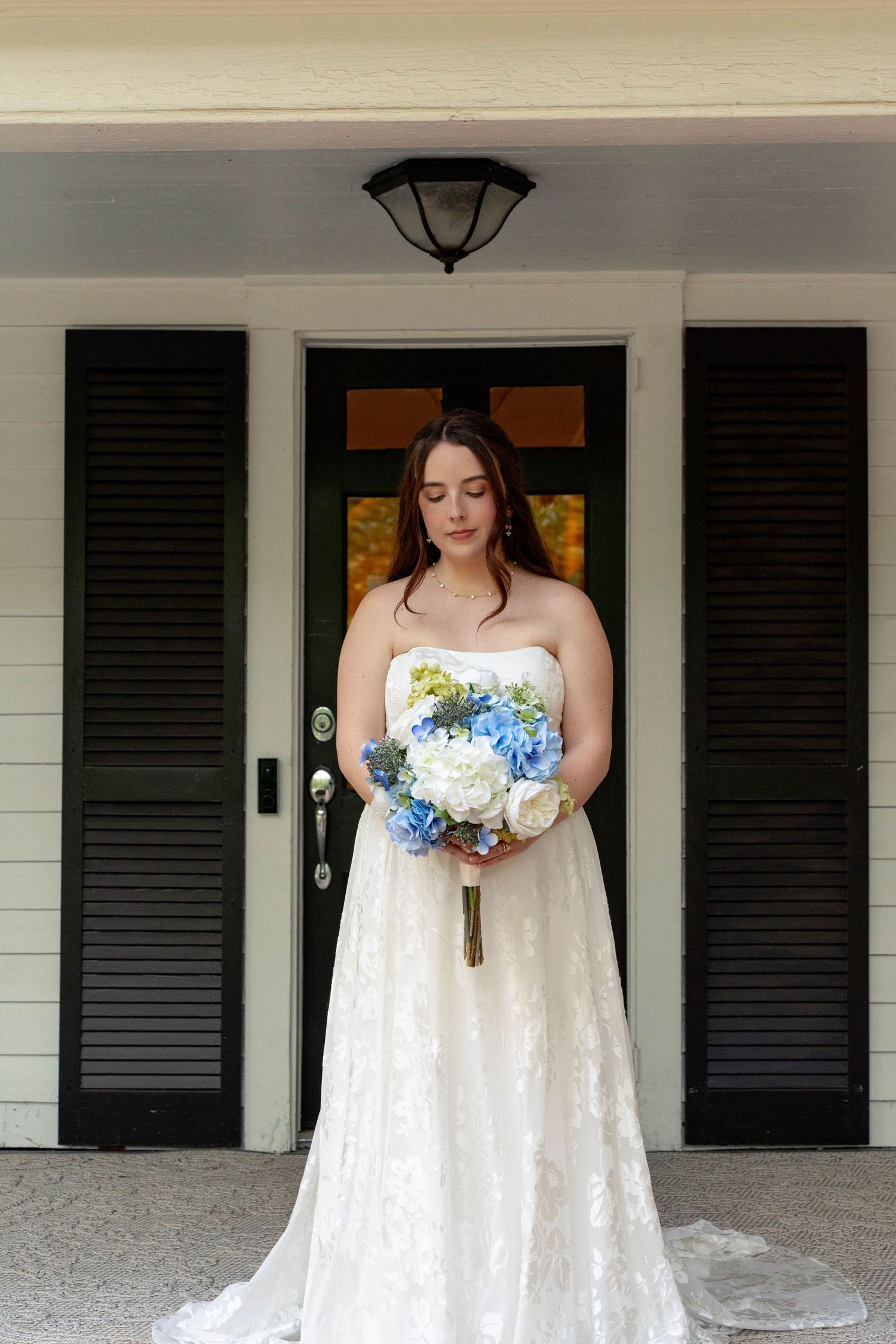 Bride in strapless lace gown holding blue and white bouquet standing before black doors.