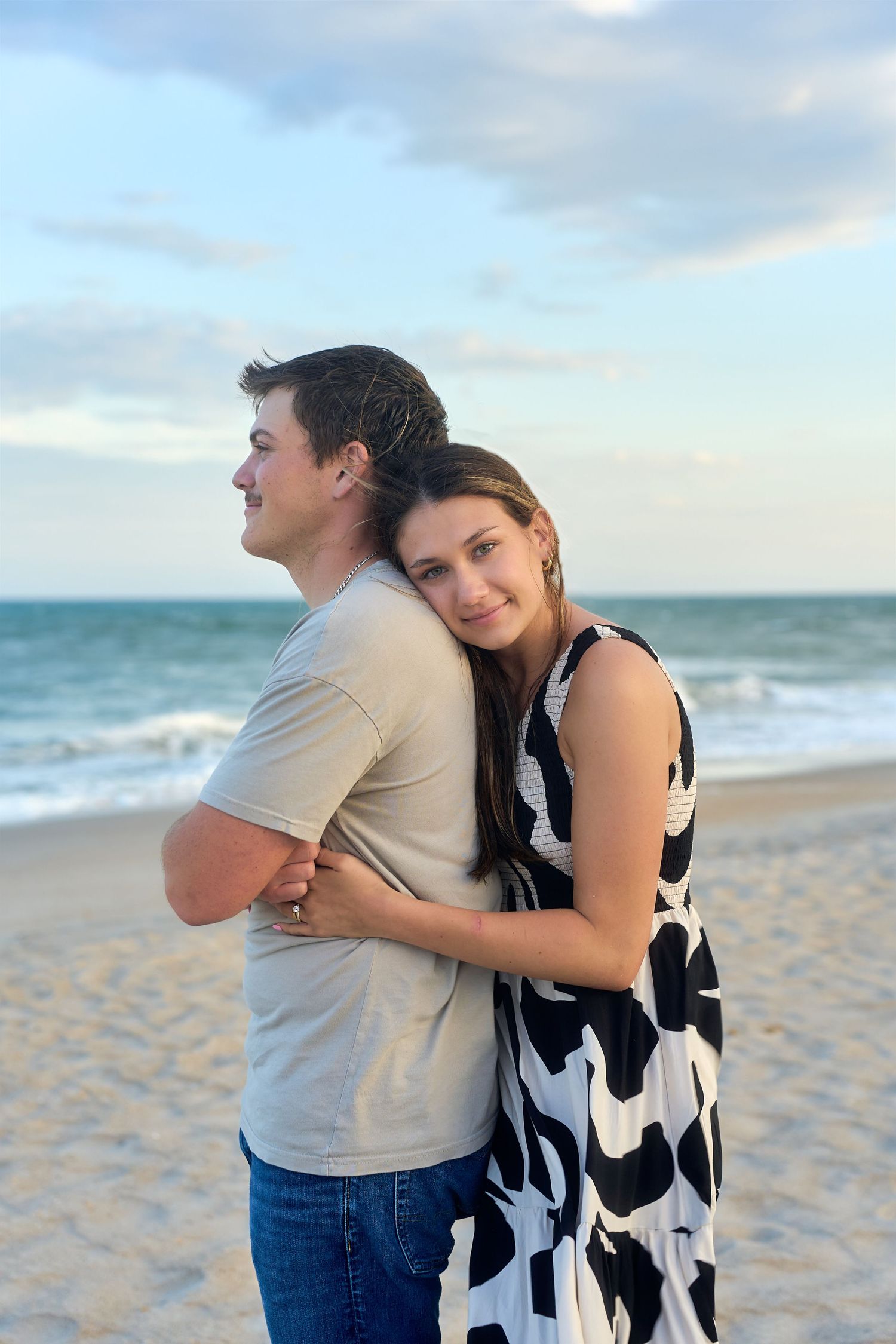 A couple embraces while standing back-to-back on a sandy beach at sunset with ocean waves in the background.