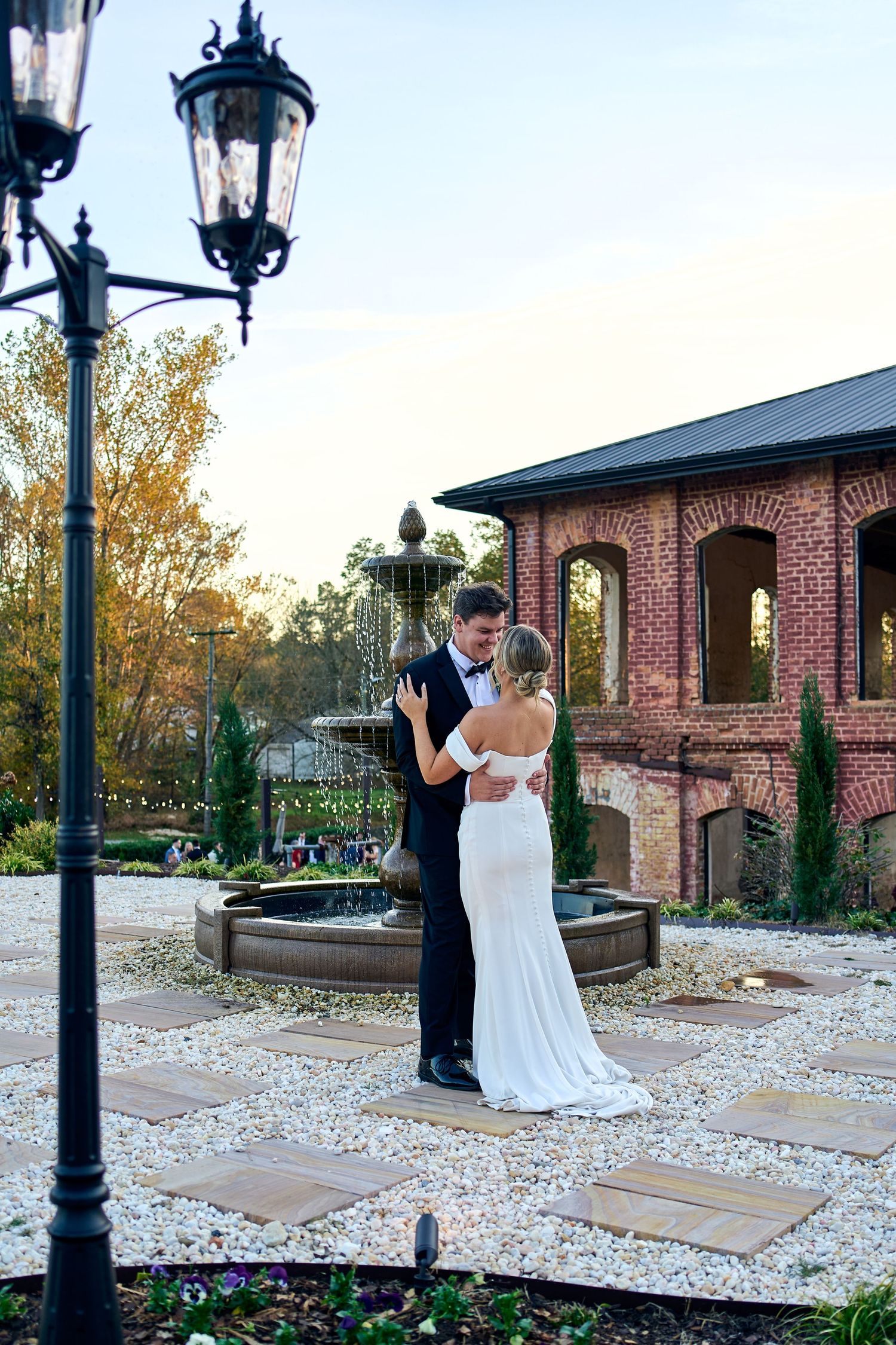 A couple shares a romantic moment on a patio by a fountain near a brick building with a vintage lamppost at sunset.