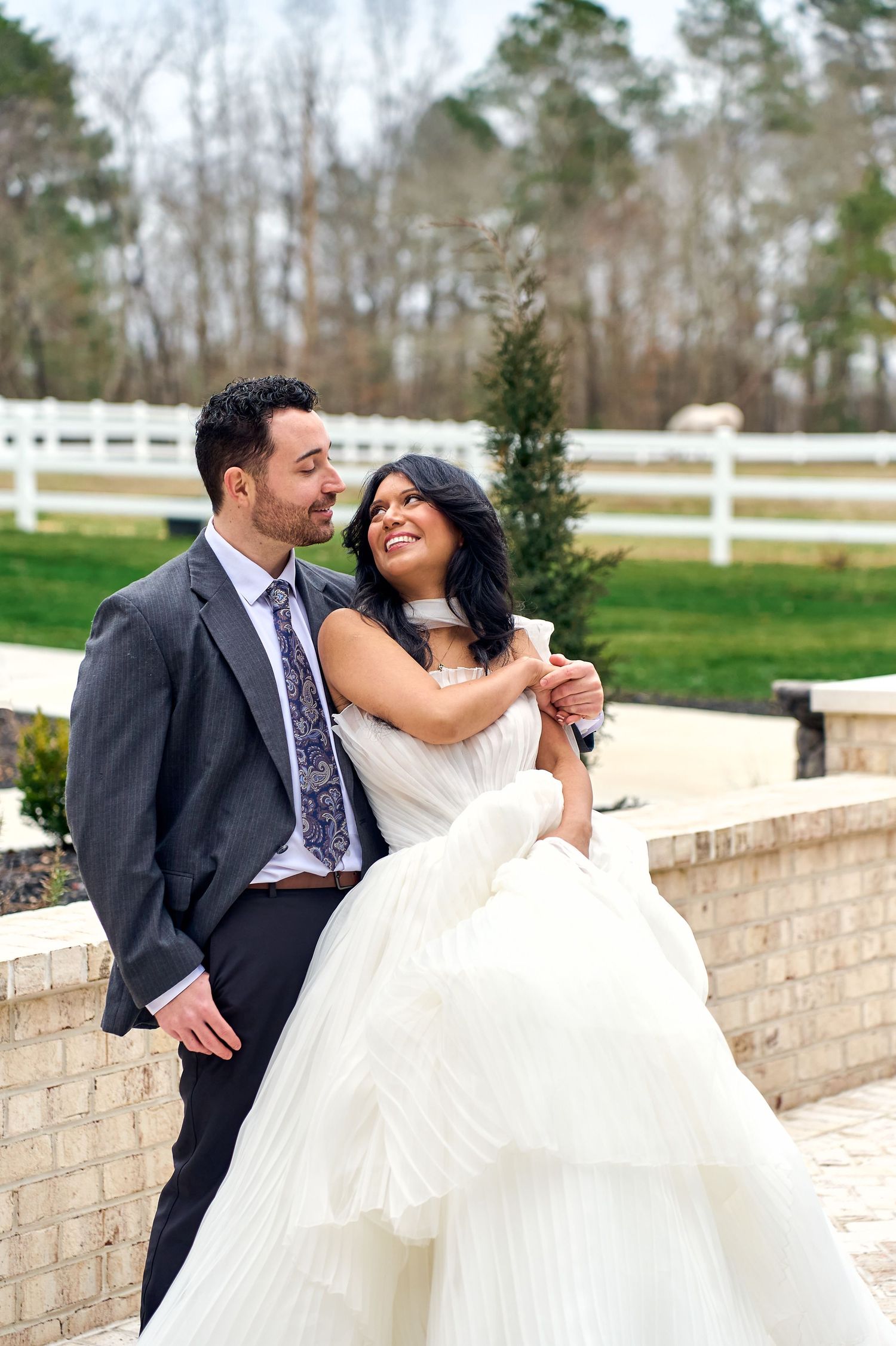 A romantic wedding couple shares a tender moment along a white brick wall and fence on their special day.