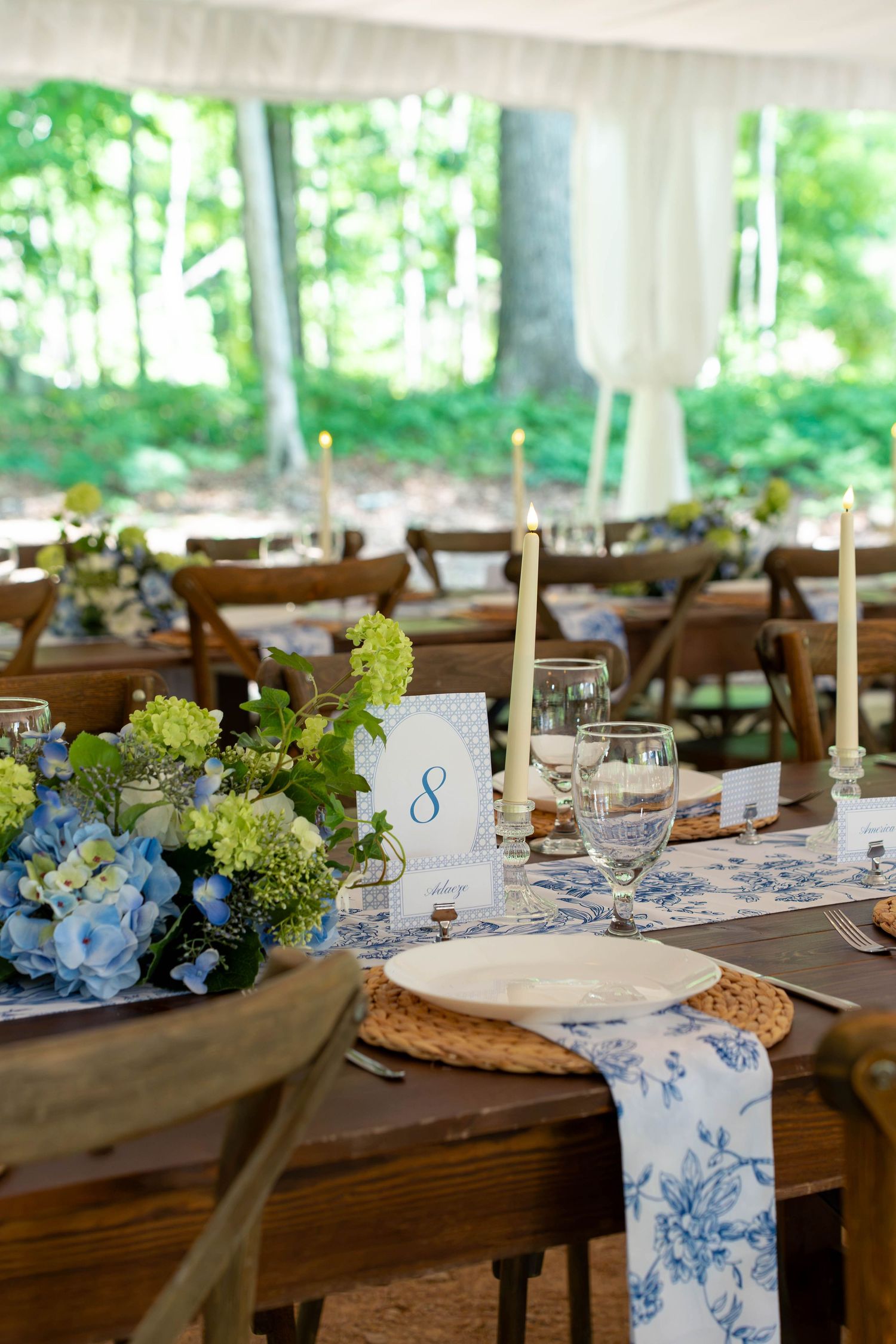 Elegant wedding reception table set with blue toile runner, hydrangeas, and candles under tent.