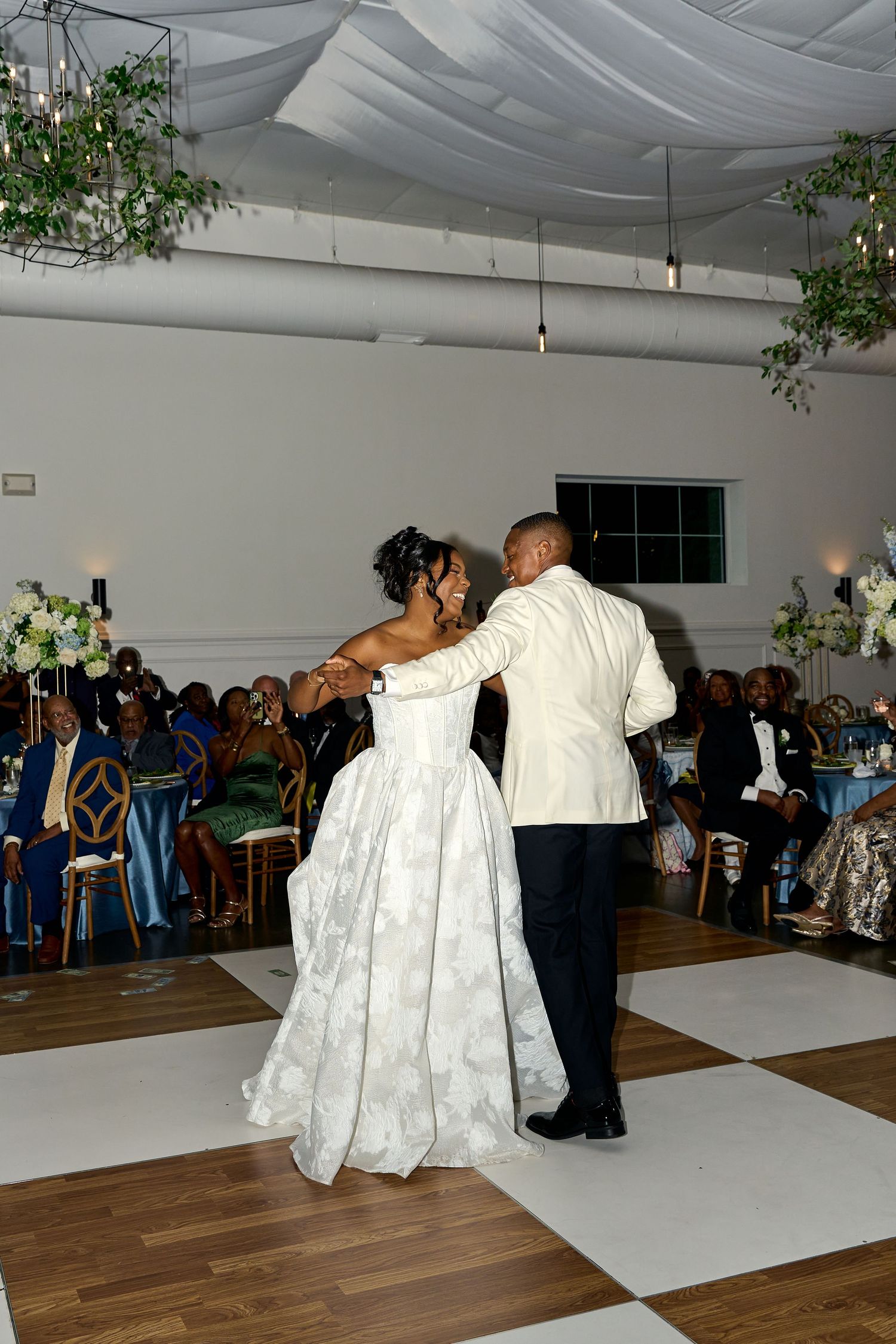 Bride and groom share first dance on checkered floor while wedding guests watch in decorated venue.