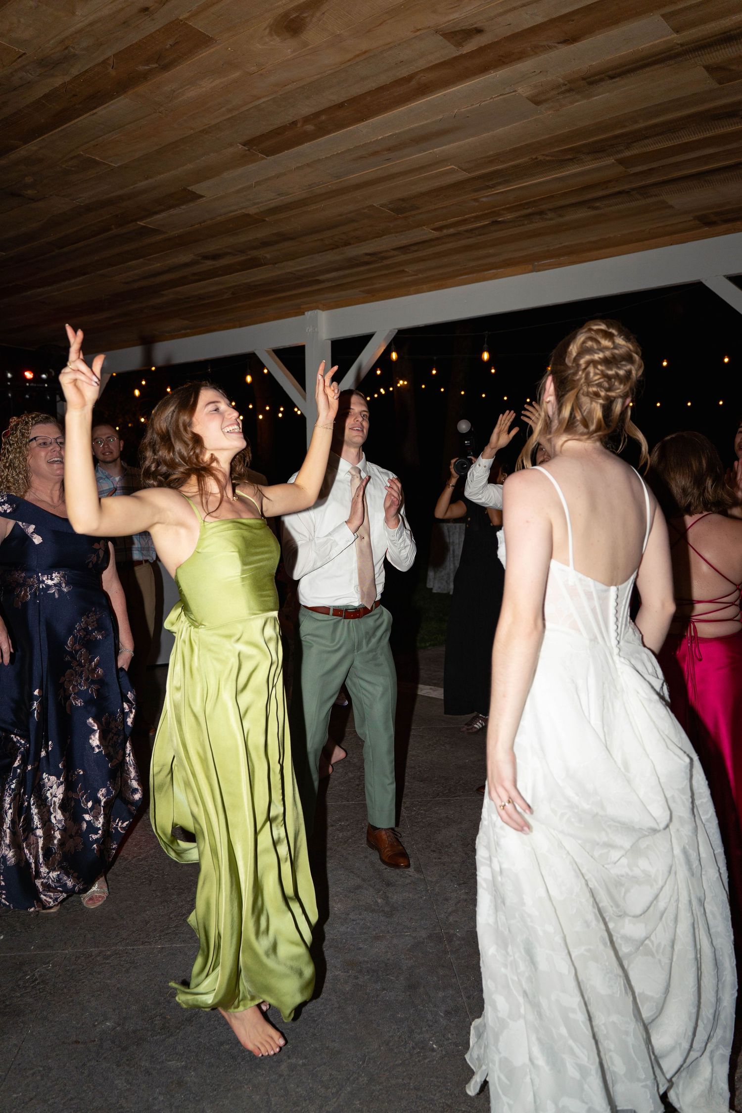 Bride and bridesmaid in green dress dancing joyfully at an outdoor wedding reception at night.