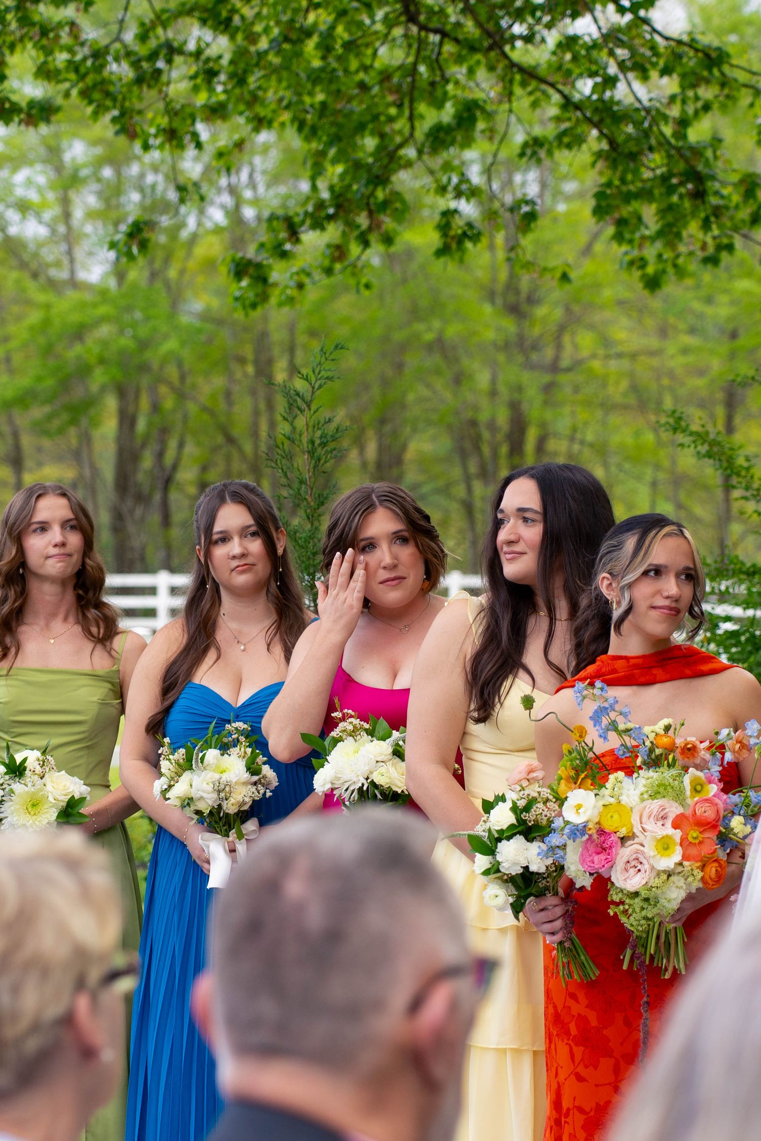 Bridesmaids in colorful gowns hold bouquets with emotional expressions during outdoor wedding ceremony.