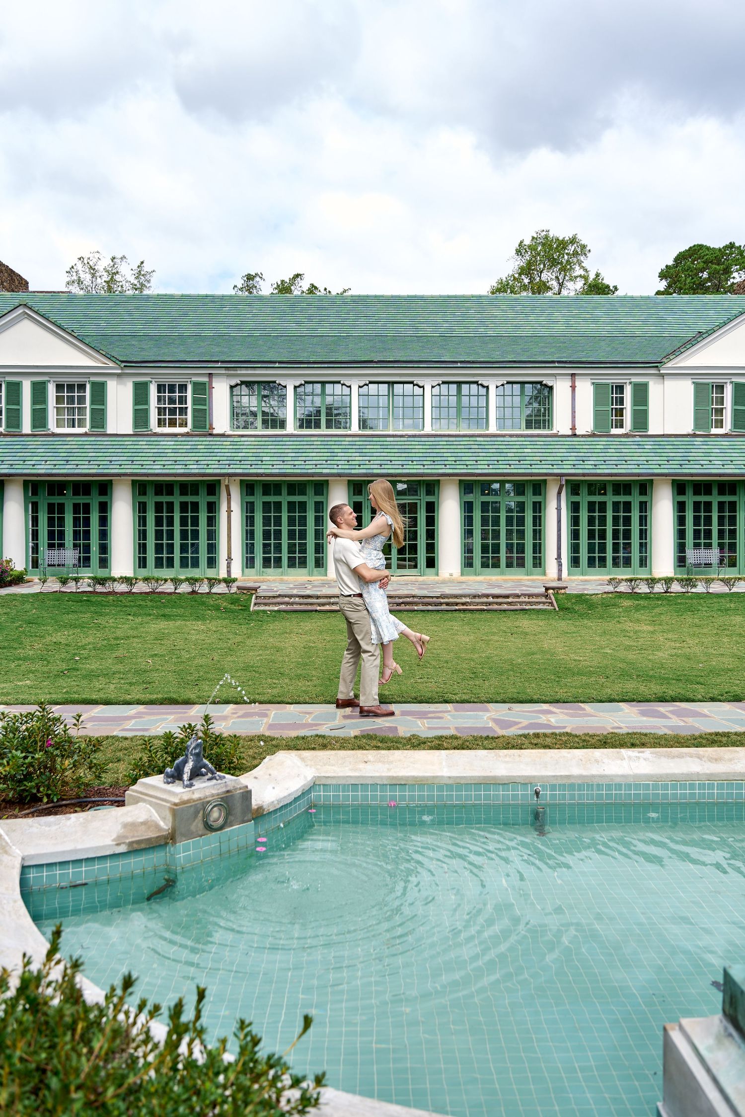 Man lifting woman by a pool in front of a large white estate with green roof.