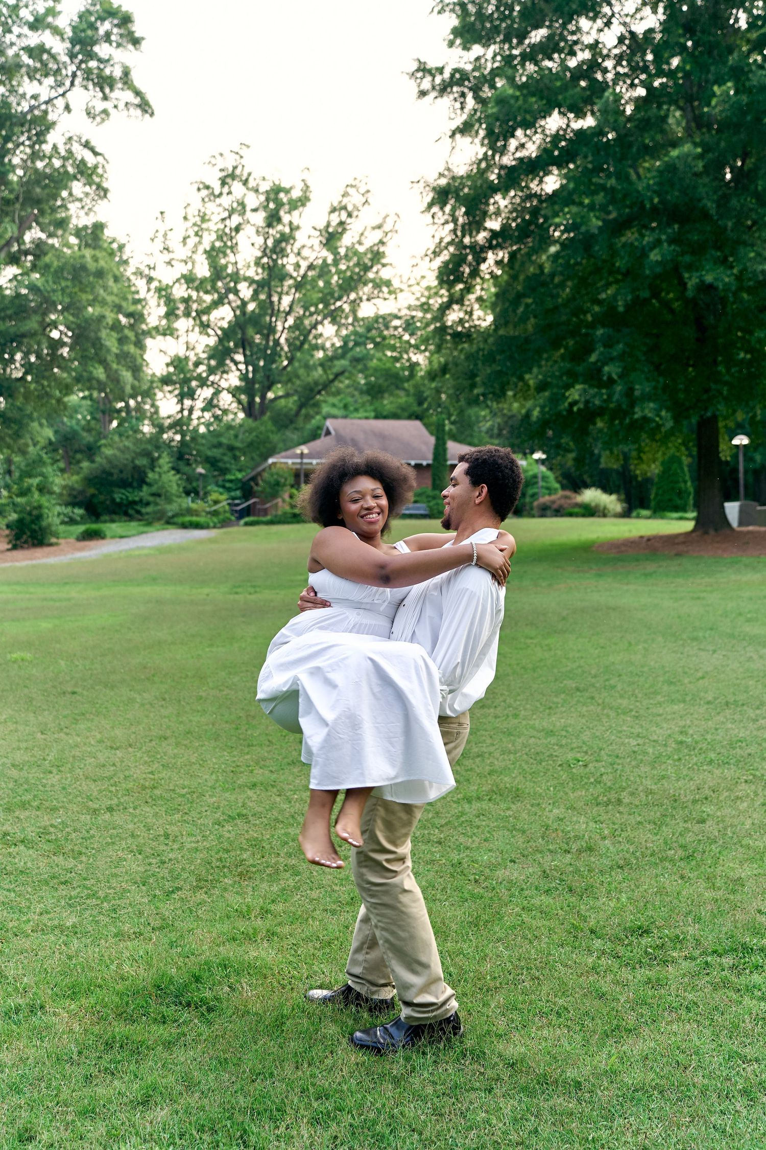 A playful couple dances and spins together on a grassy lawn surrounded by trees.