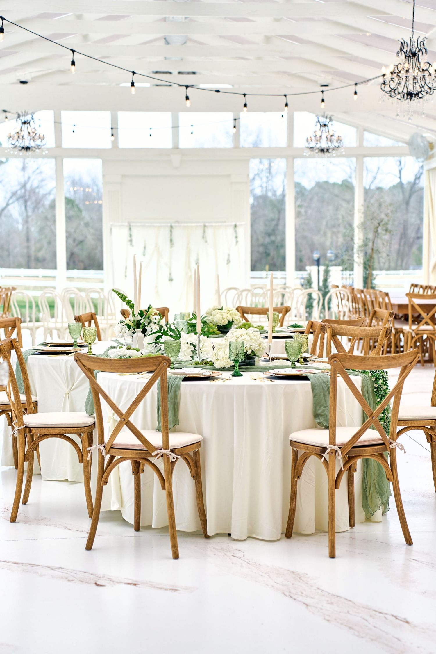 Elegant wedding reception table with white linens, wooden crossback chairs, and greenery centerpieces in a bright sunlit venue.