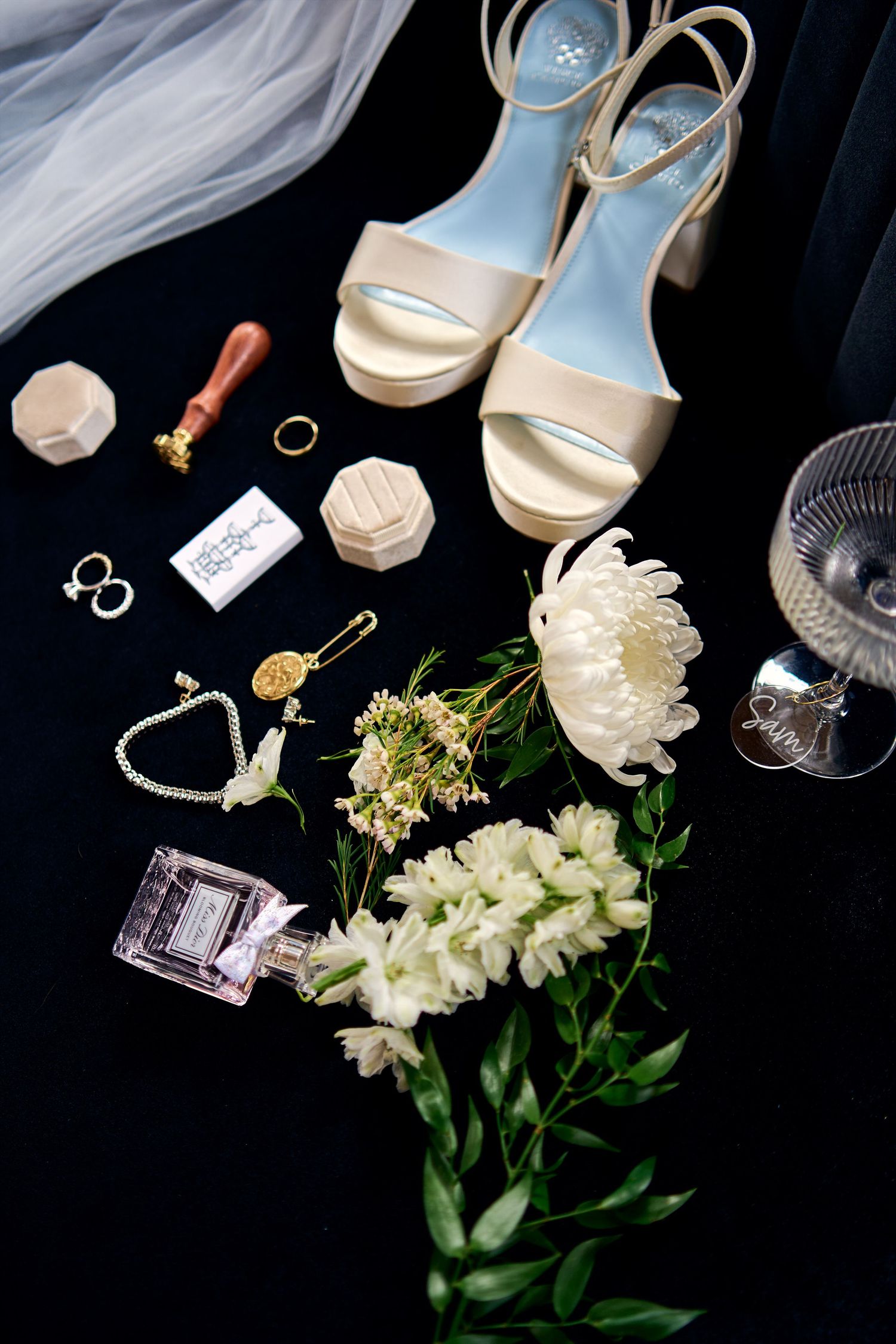 Wedding accessories flatlay with gold sandals, white flowers, rings, perfume and other bridal details on black background.