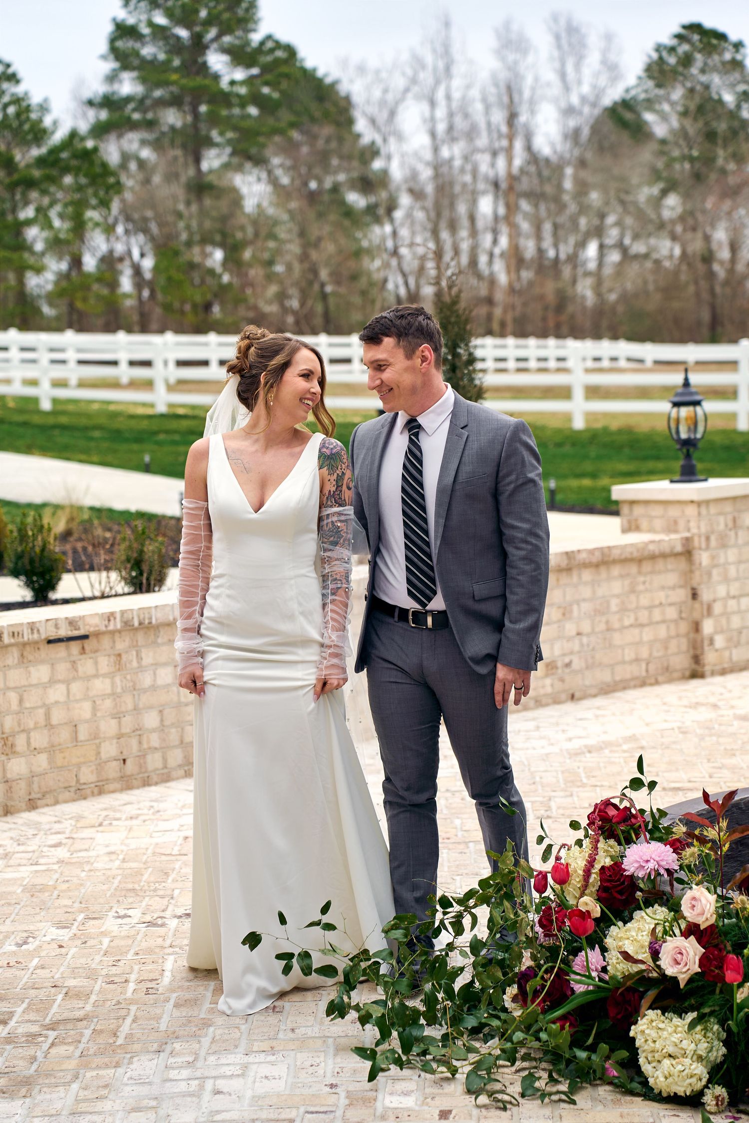 A romantic couple dressed in wedding attire stand together on a brick path by colorful floral arrangements and a white fence.