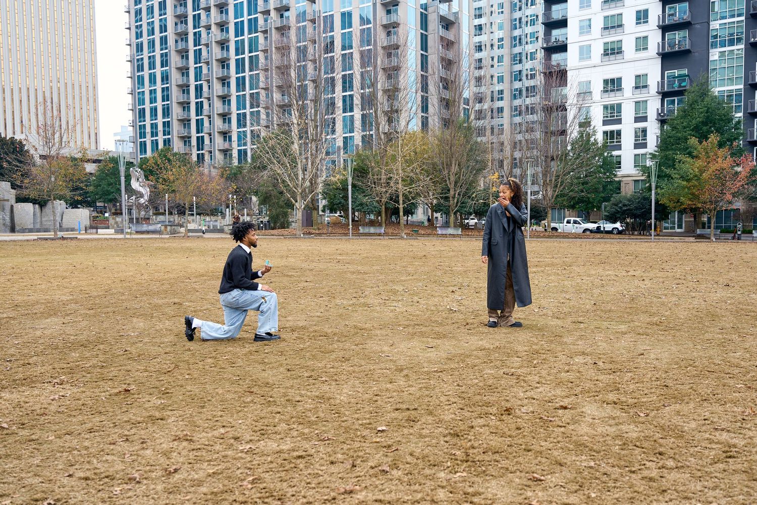 Man proposing on one knee to woman in urban park with city skyscrapers in background. North Carolina Wedding Photographer.
