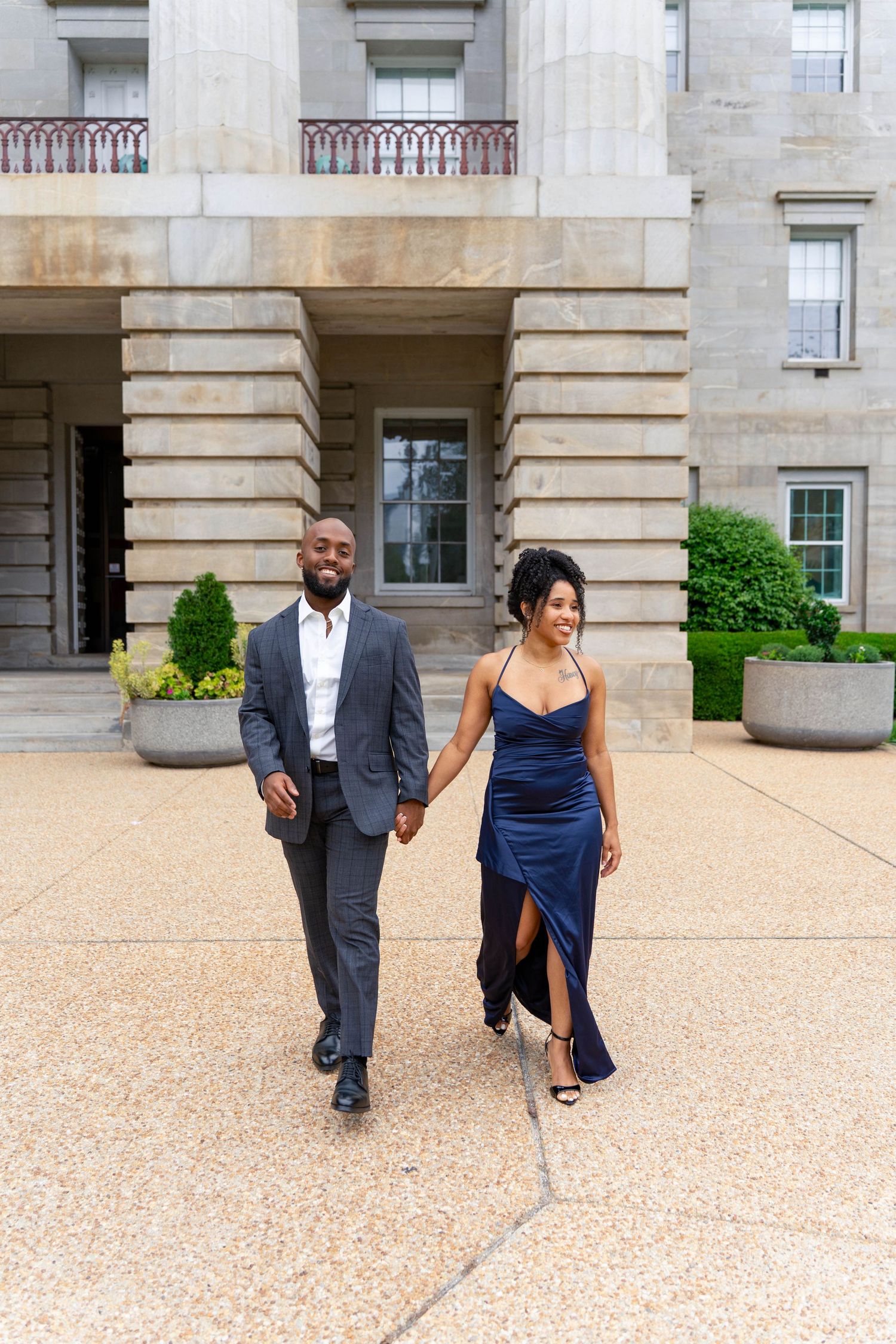 Couple in formal attire walk hand in hand in front of a grand stone building.