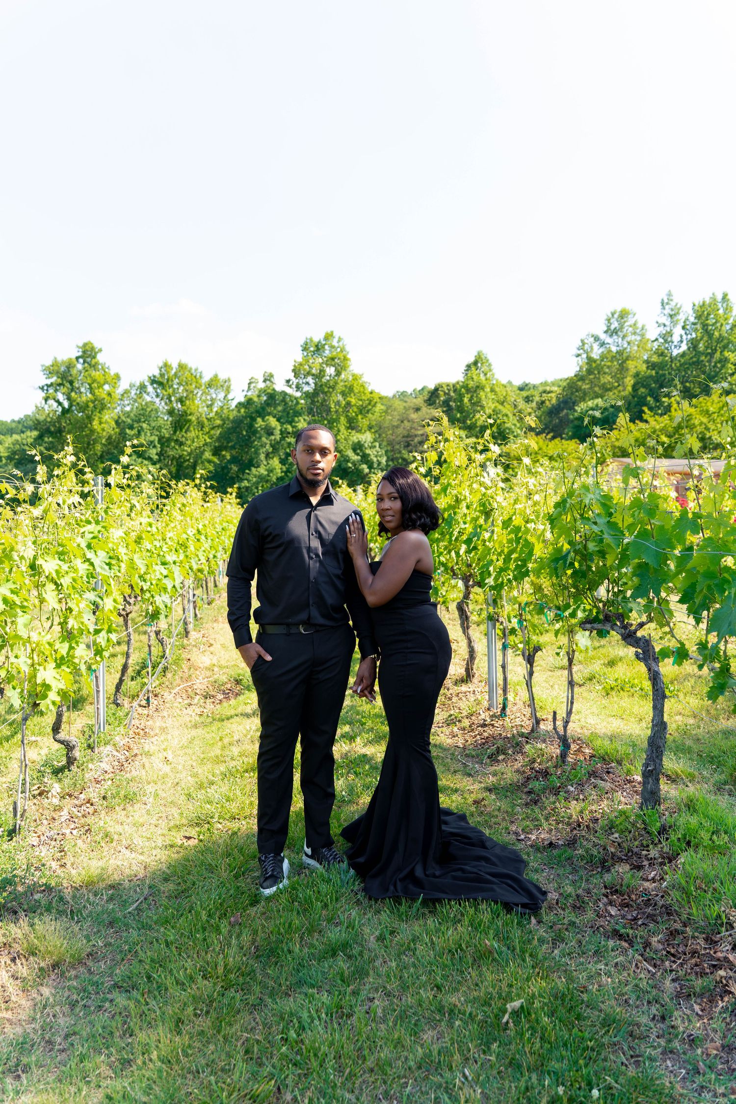 Couple in coordinating black formal wear posing together among lush green vineyard rows outdoors.