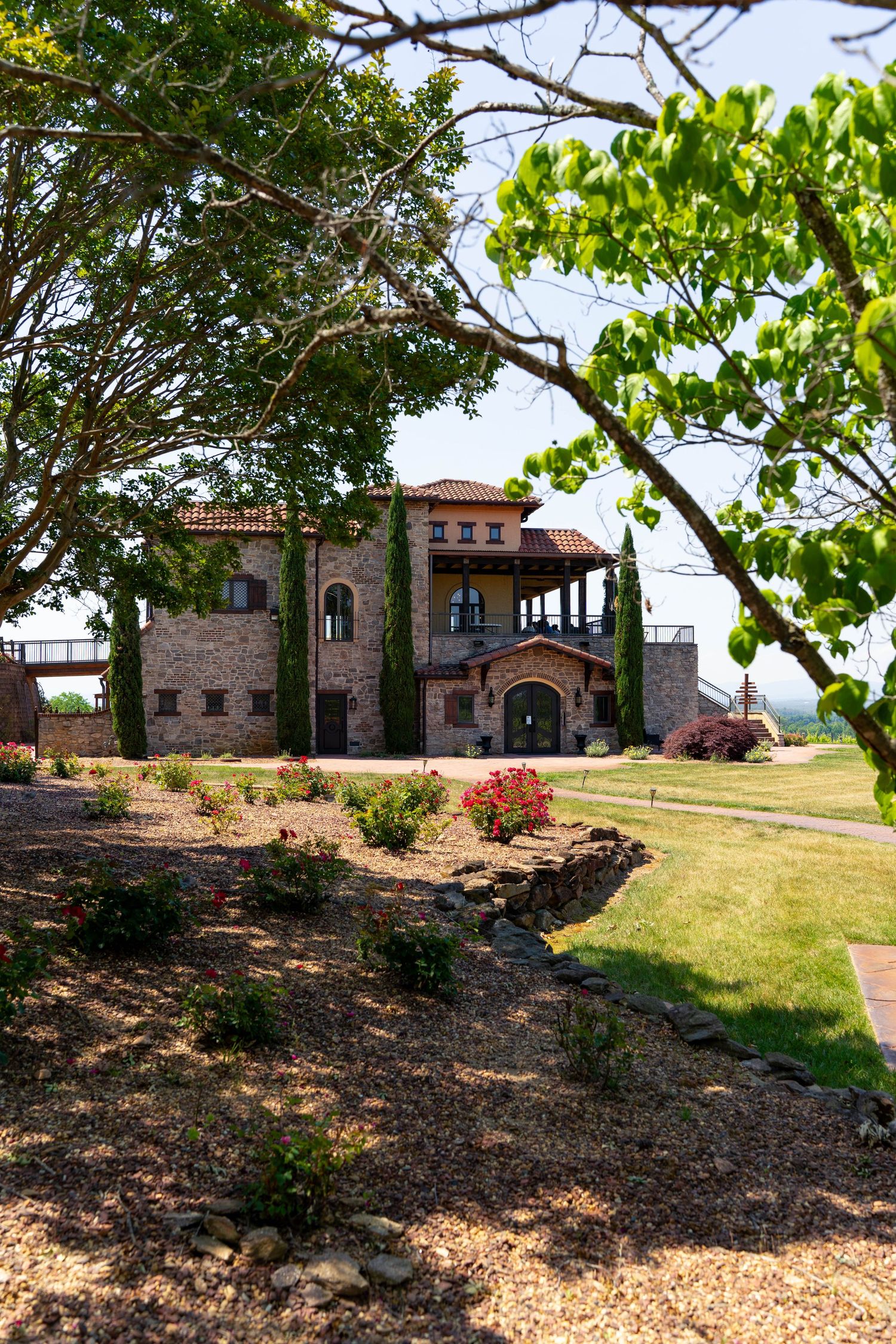 Stunning Italian-style stone mansion surrounded by lush gardens and cypress trees on a sunny day.