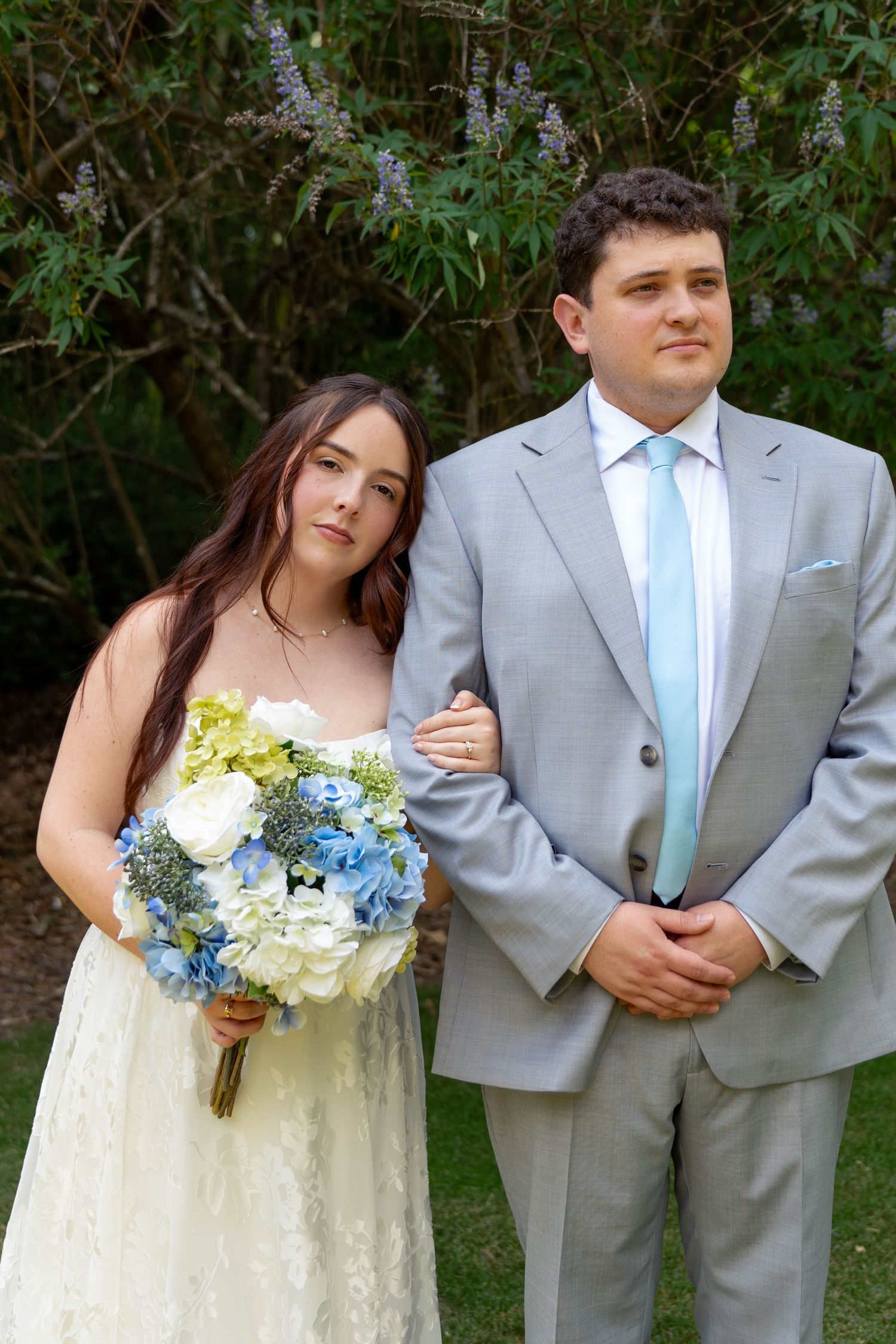Bride holding blue and white hydrangea bouquet stands with groom in gray suit and light blue tie outdoors.