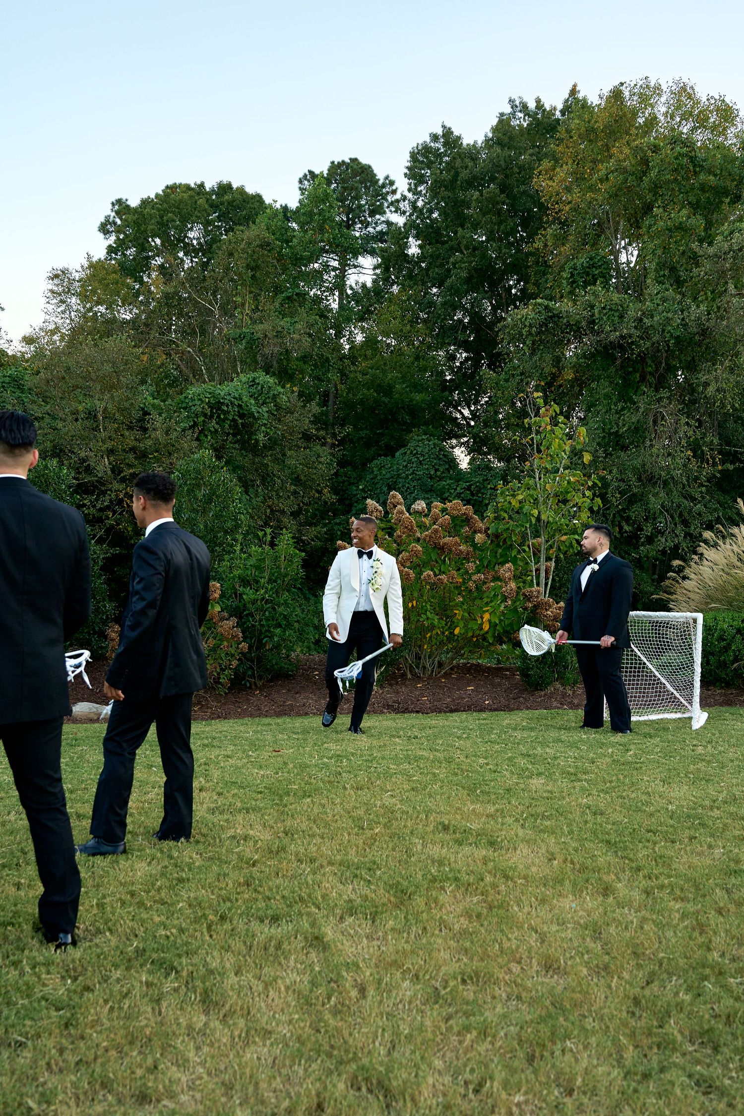 Groom in white jacket plays lawn games with groomsmen in black tuxedos at outdoor wedding reception.