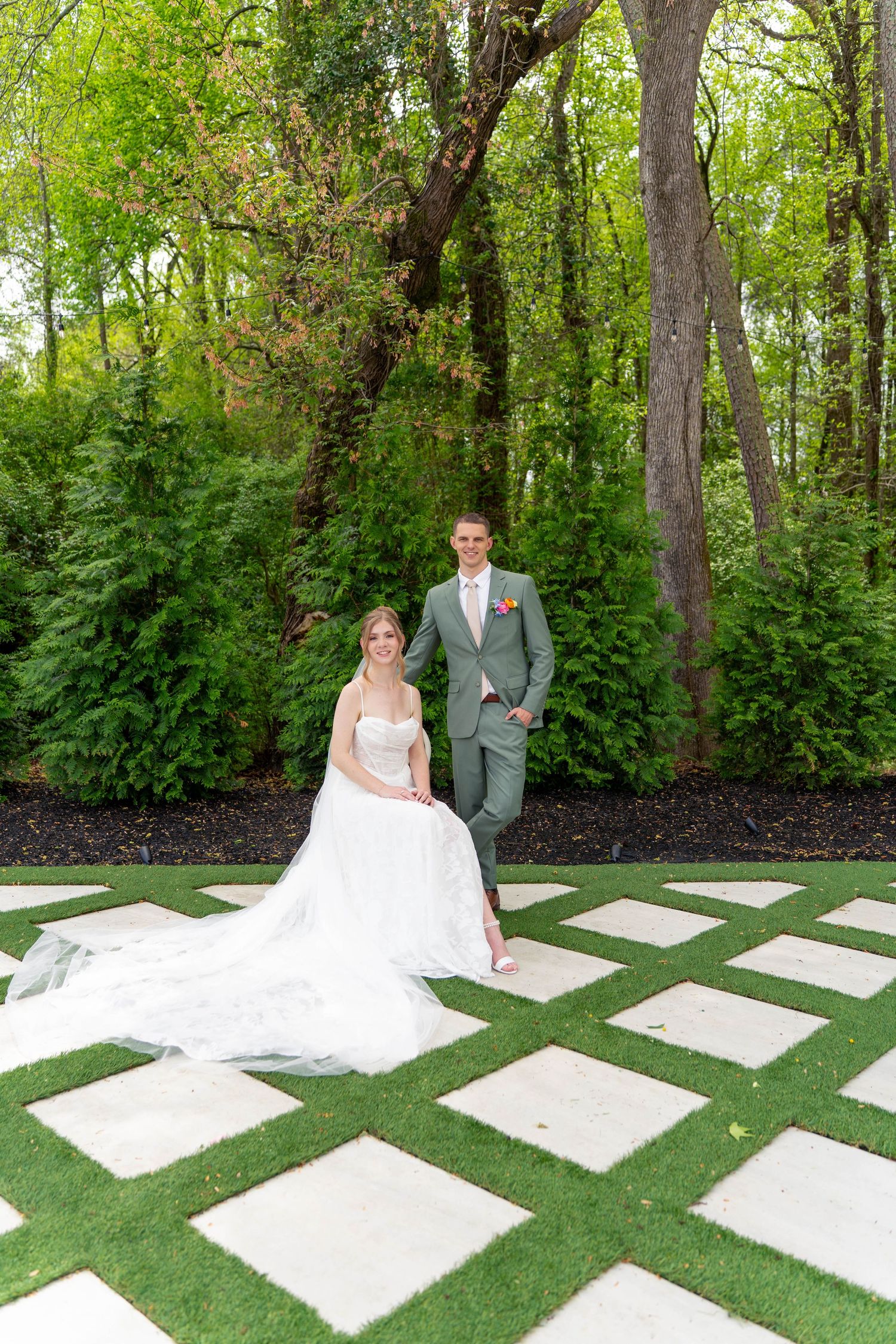 Bride in white ballgown sits on stone tile as groom in sage suit stands beside her in garden setting.