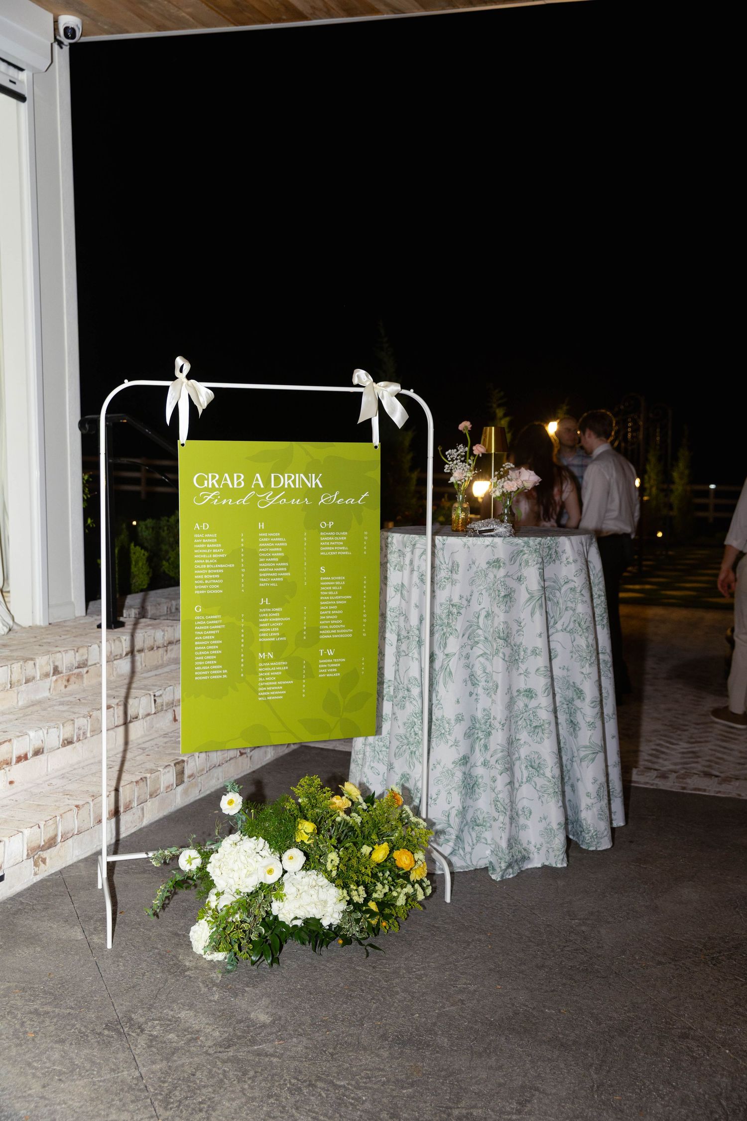 Green wedding seating chart sign on white rack with white floral arrangement at night reception.