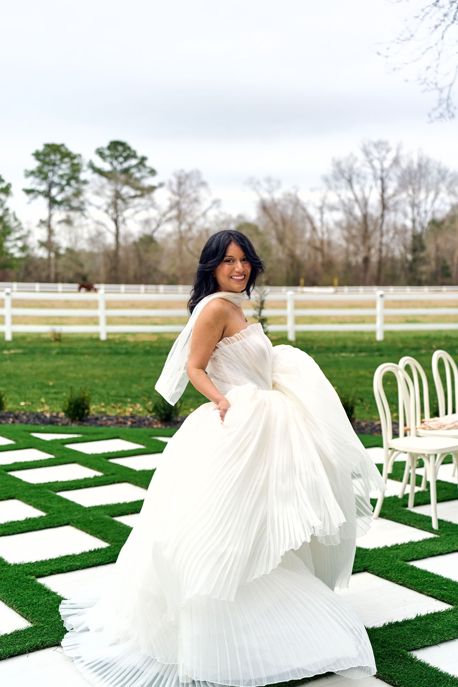 A bride in a pleated white wedding gown poses outdoors on a grass lawn with white chairs and fencing in the background.