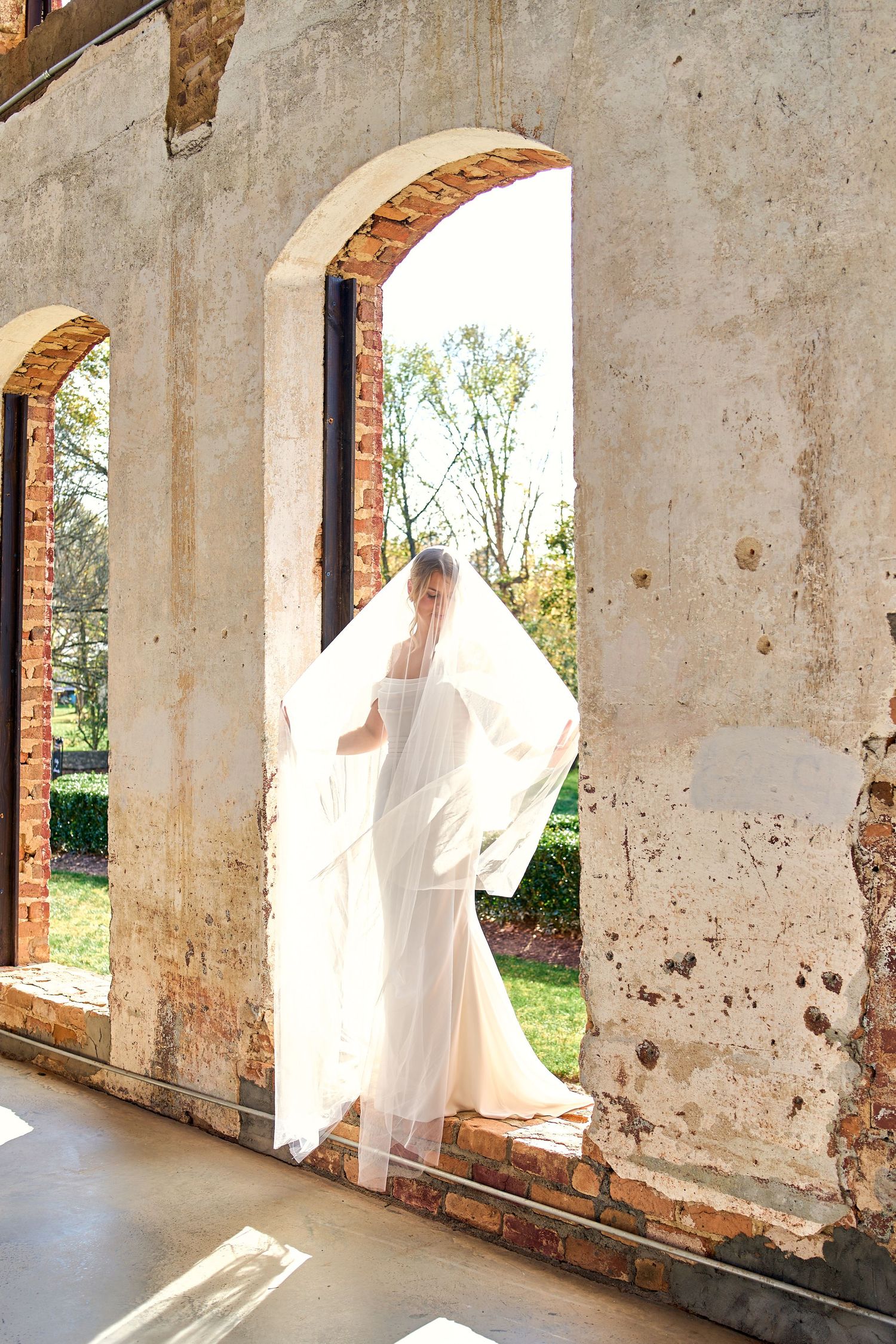 A flowing white wedding dress catches sunlight through an archway in a rustic brick and plaster building.