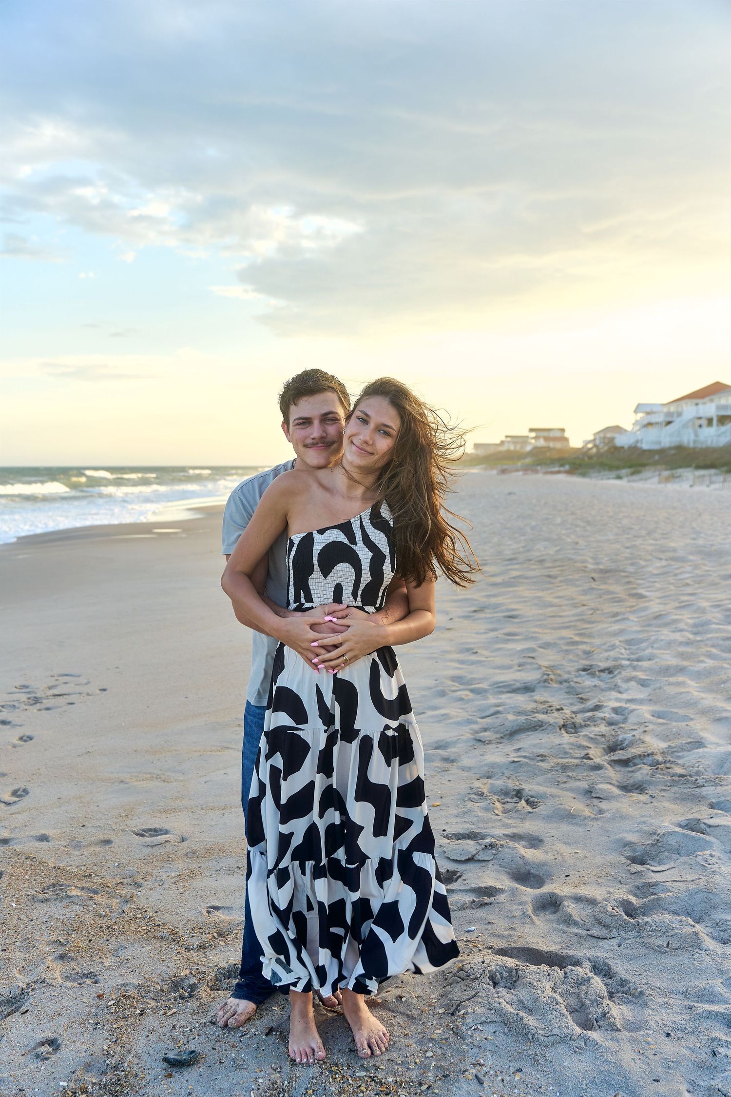 A couple embraces on a sandy beach at sunset, with waves and beach houses in the background.