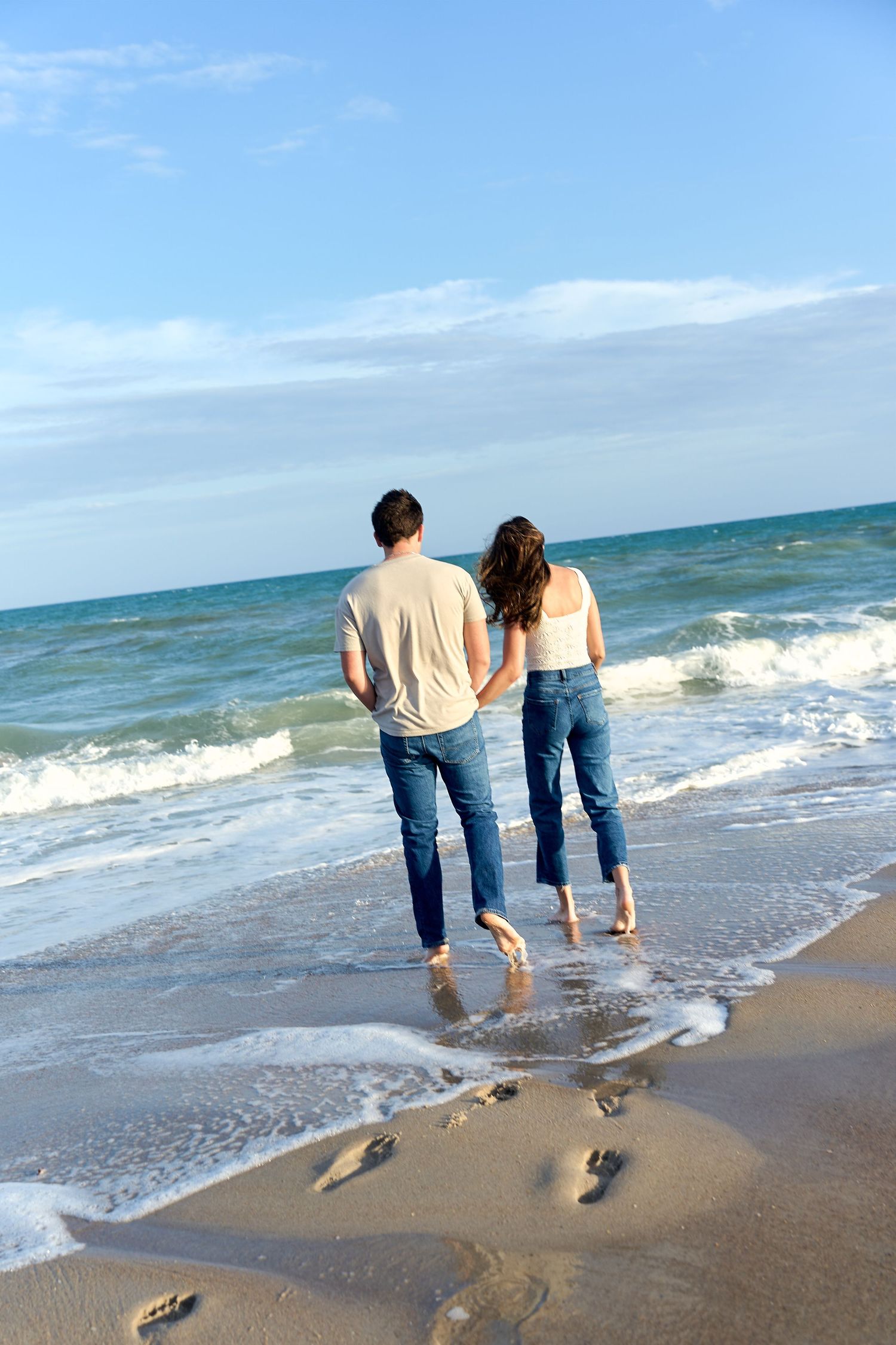 Two people in casual denim walking barefoot along the beach shoreline leaving footprints in the wet sand.
