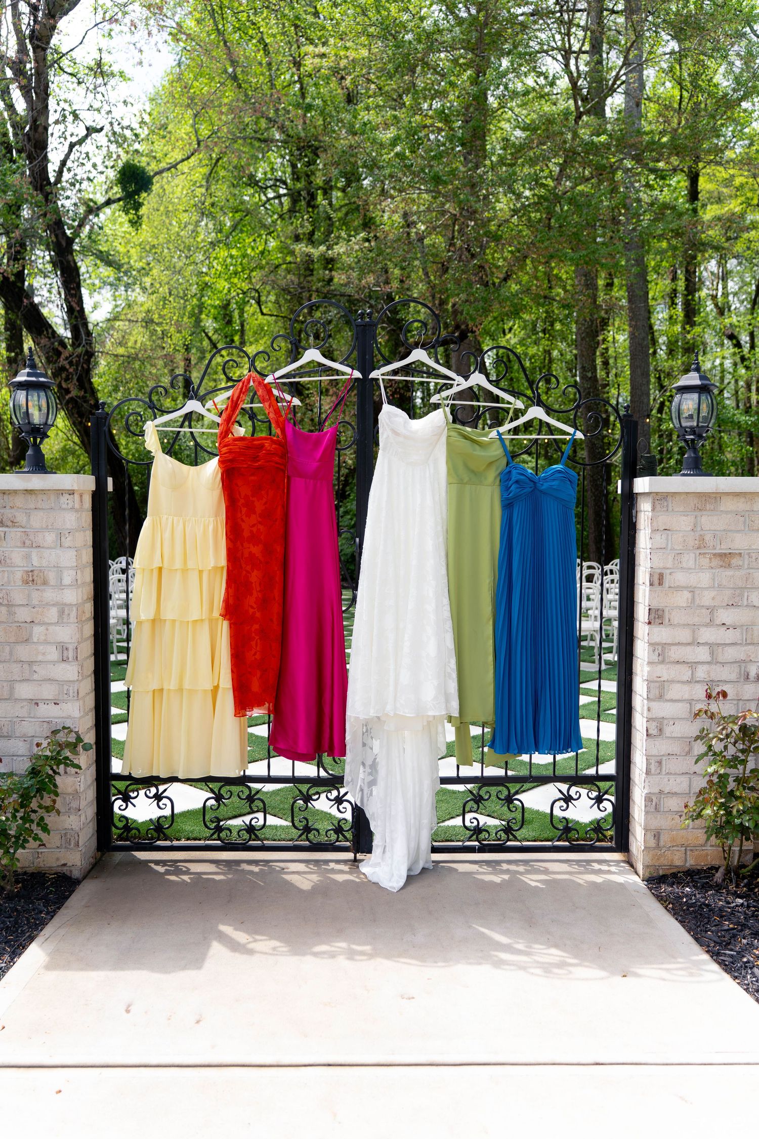 Colorful bridesmaid dresses and white wedding gown hanging on ornate iron gate surrounded by trees.