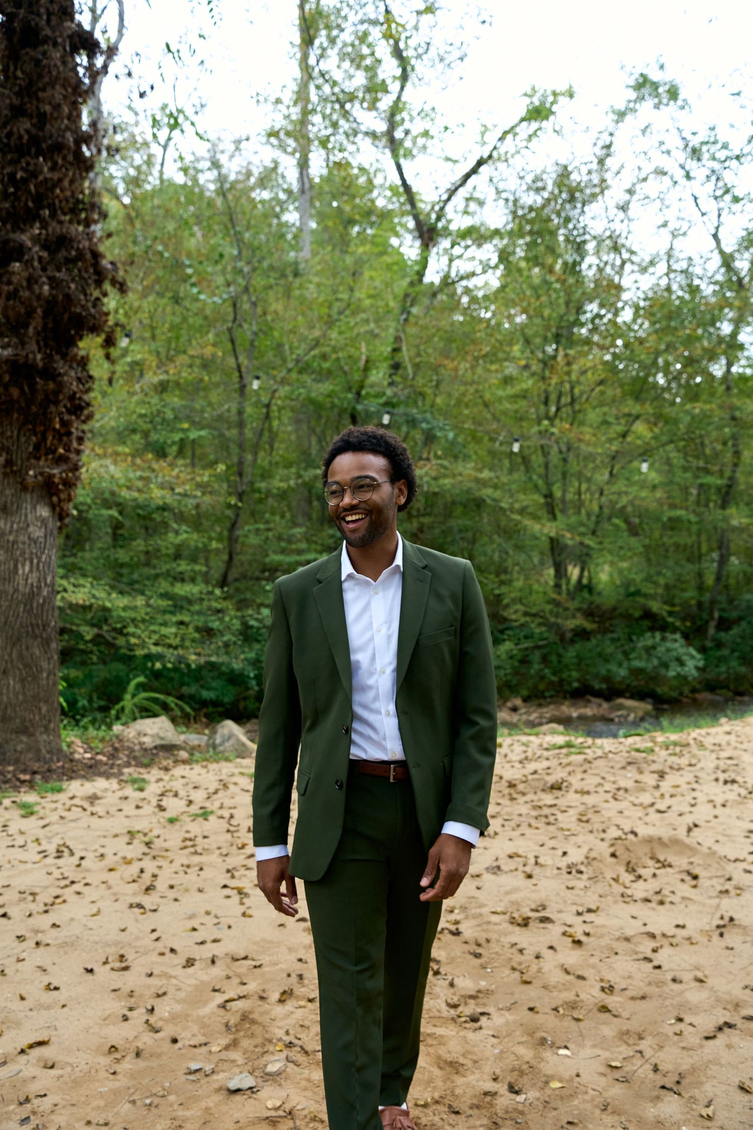 Smiling young Black man in a green suit laughing while standing on a sandy area surrounded by trees.
