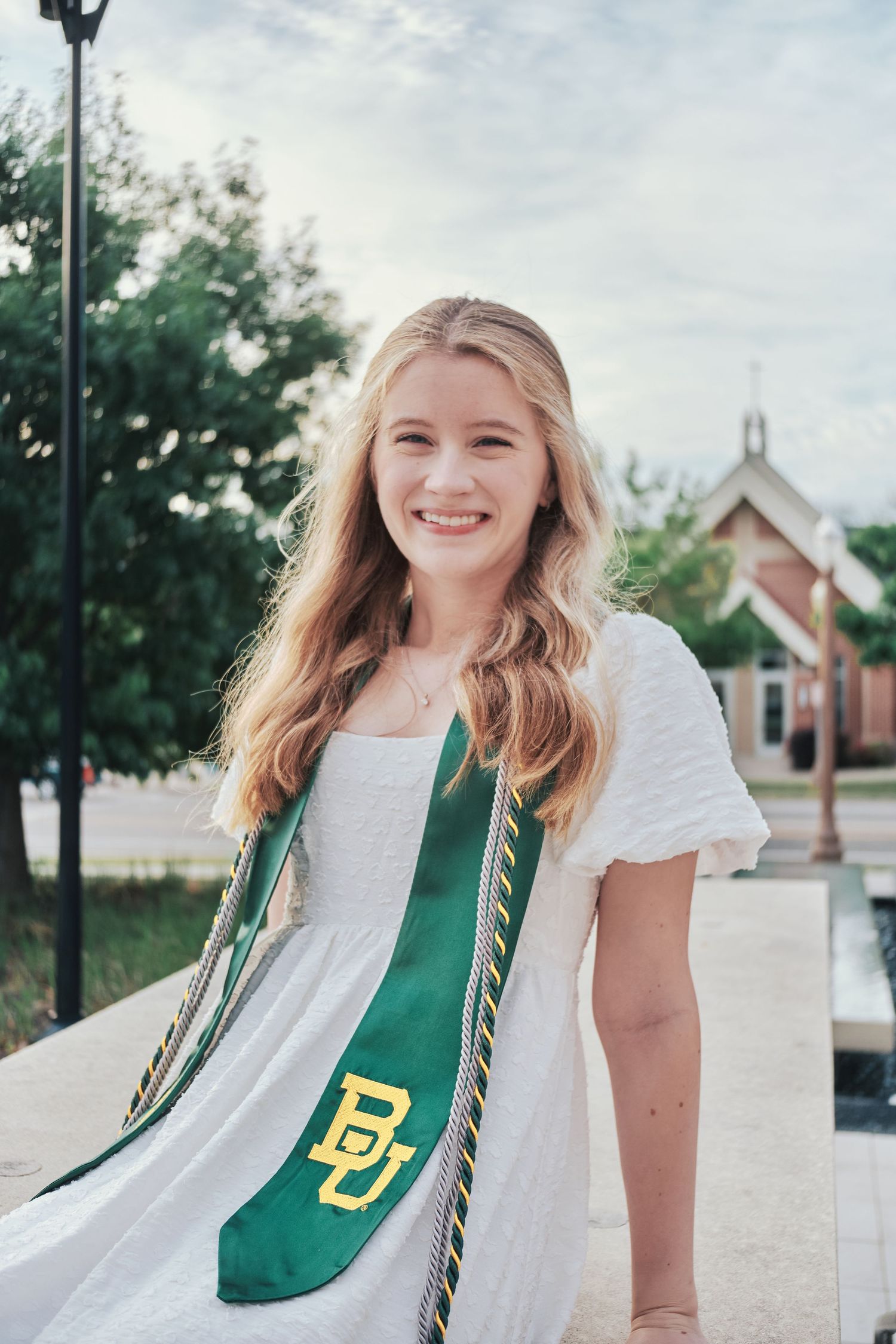 Smiling blonde graduate wearing a Baylor University green and gold sash in a white dress outdoors.