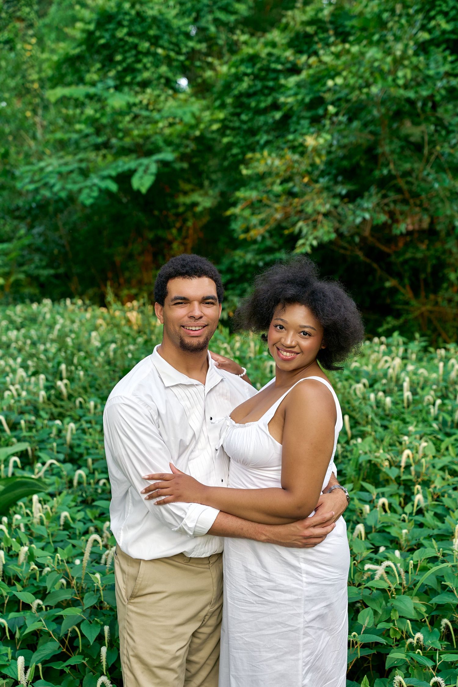 Couple in white attire embraces lovingly in a lush green garden setting filled with foliage.