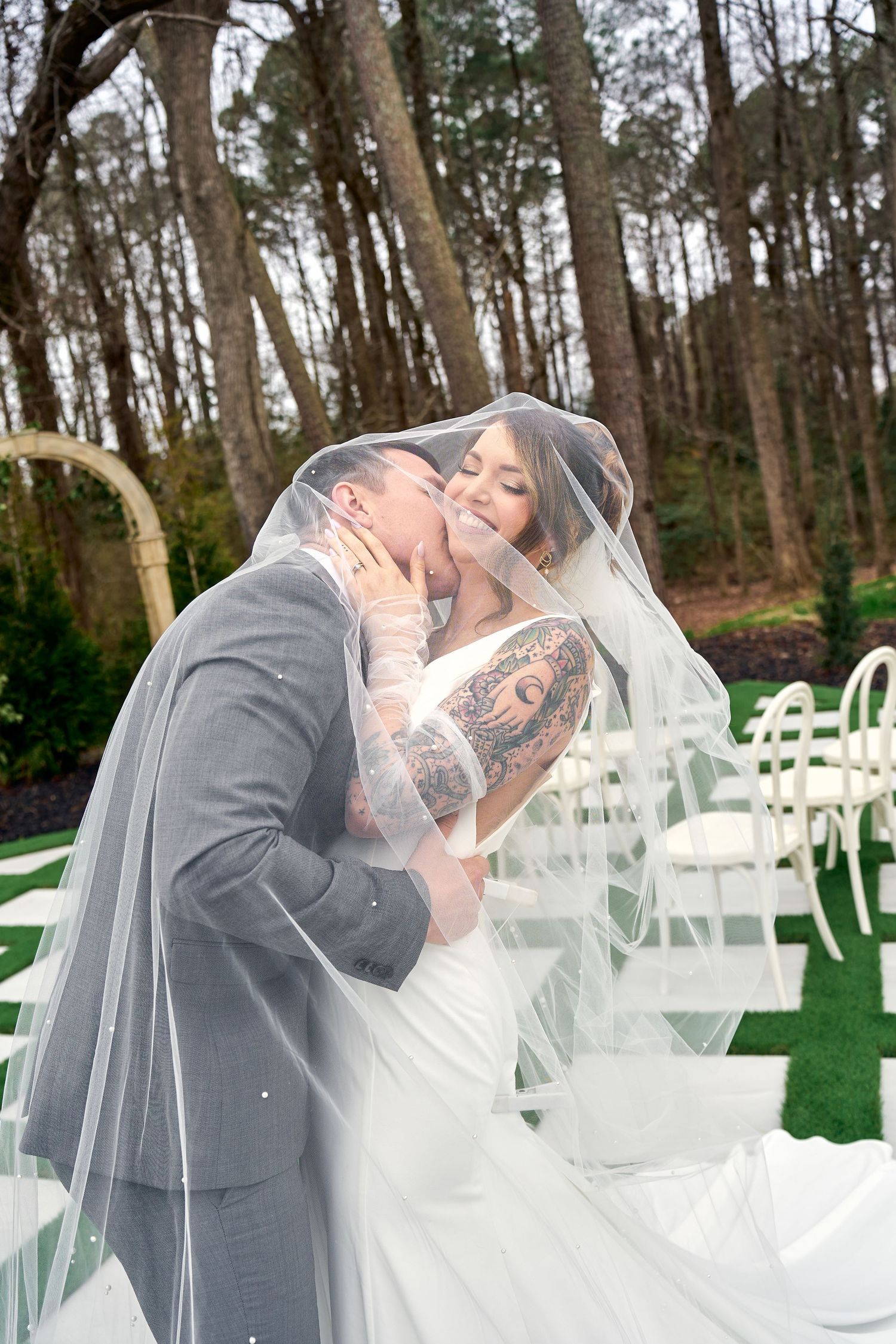 A romantic outdoor wedding ceremony with white chairs and forest backdrop showing newlyweds sharing a tender kiss.