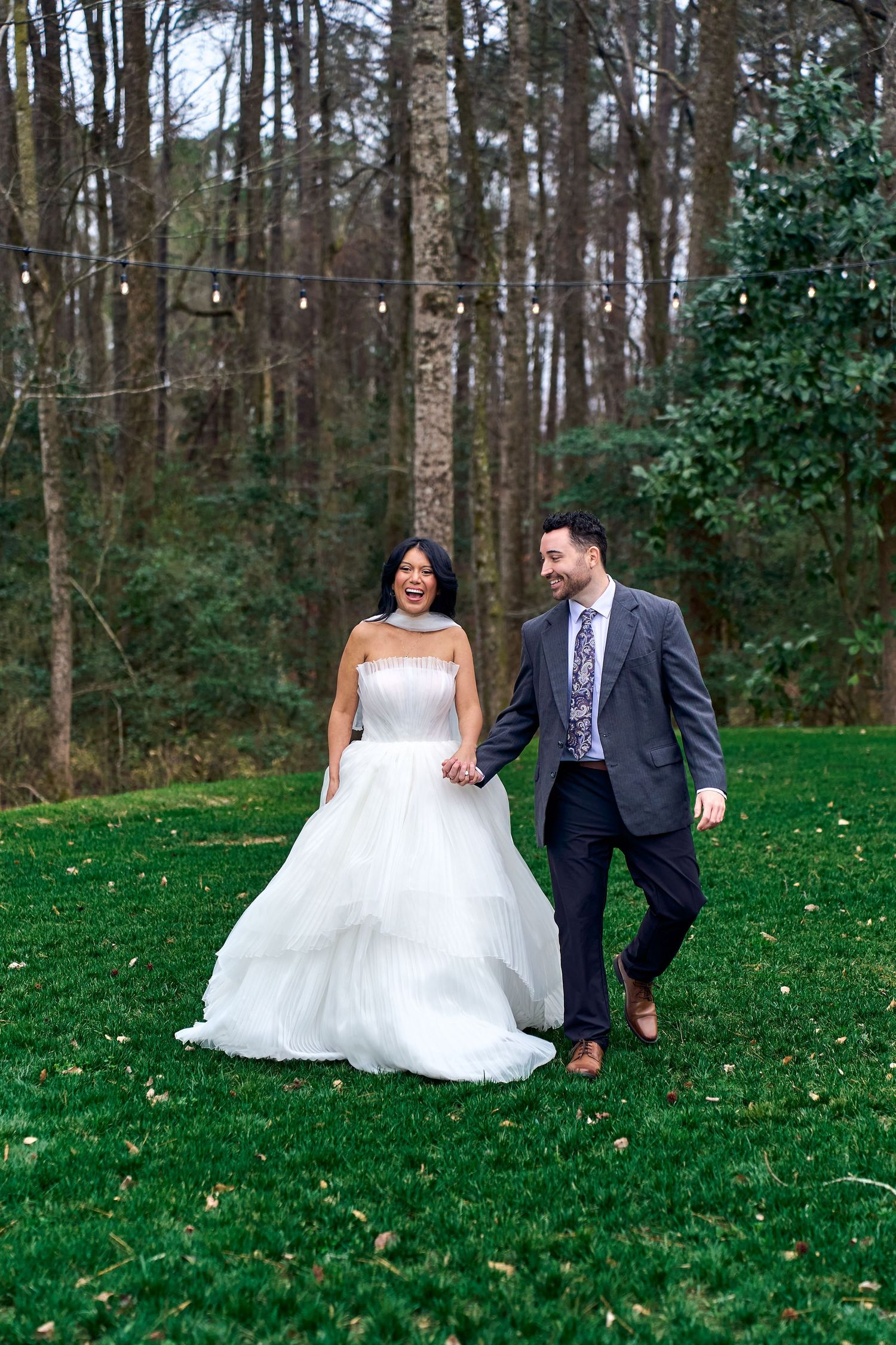 A couple in wedding attire walks hand in hand across a lush green lawn surrounded by tall trees and string lights.