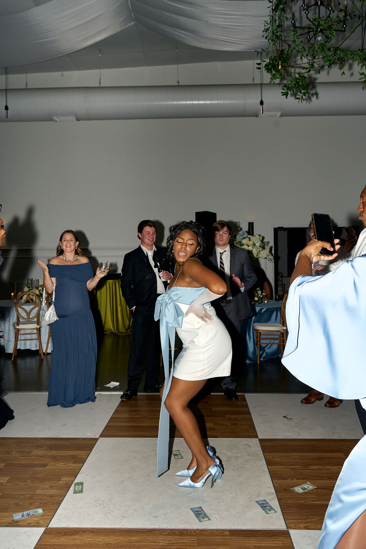 Wedding guest in white mini dress with blue bow dances on the checkered floor at reception.