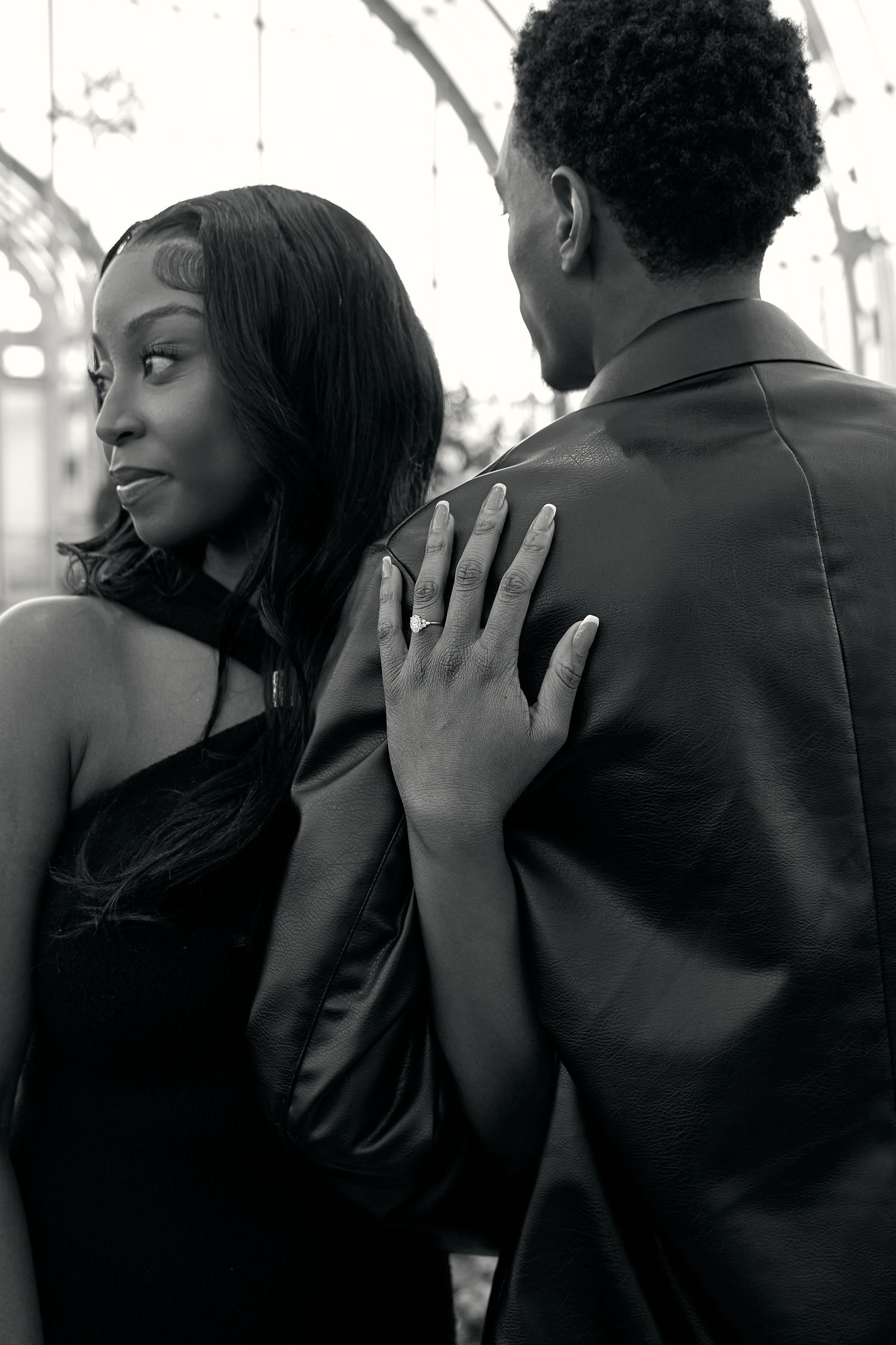 Black and white photo of an engaged couple in elegant black outfits, woman showing diamond ring while touching mans shoulder.