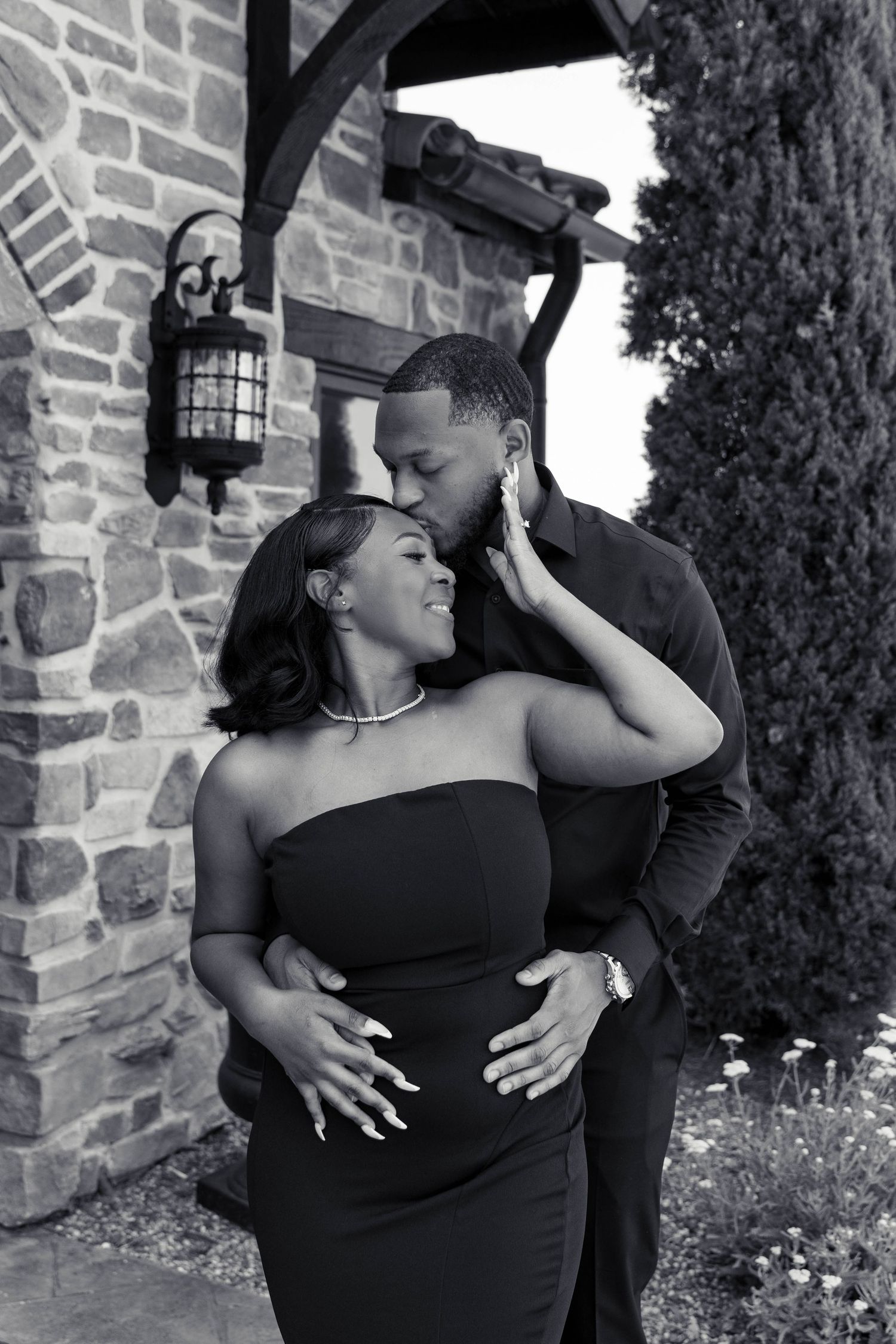 Black and white romantic photo of couple embracing and kissing near stone arch building during engagement session.